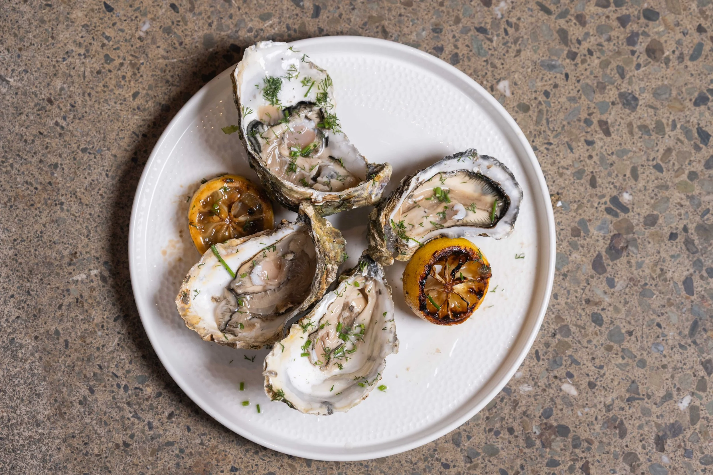 A white plate with six oysters on the half shell garnished with chopped herbs, along with two grilled lemon halves on a speckled brown surface.