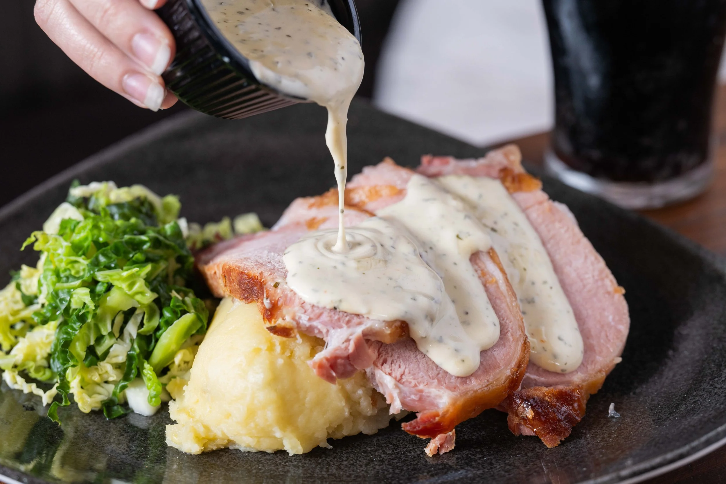 A plate of mashed potatoes with slices of pork roast covered in white gravy, served with a side of cooked greens, and a glass of dark soda in the background.