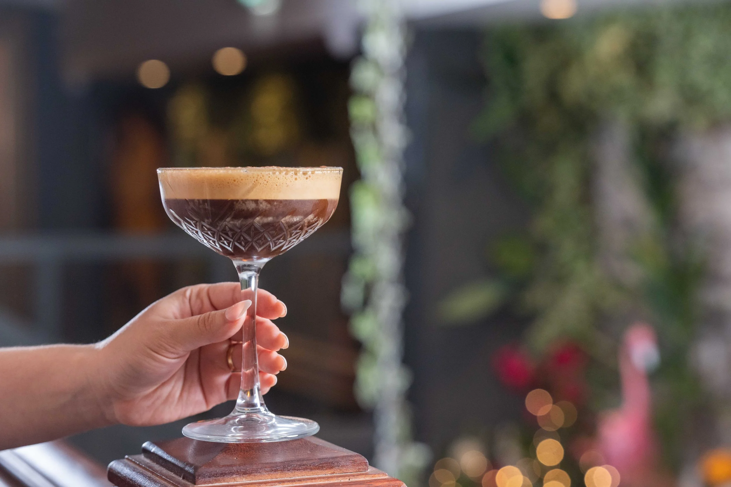 A hand holding a coupe glass filled with an espresso martini cocktail, with a blurred background featuring greenery and warm lighting.