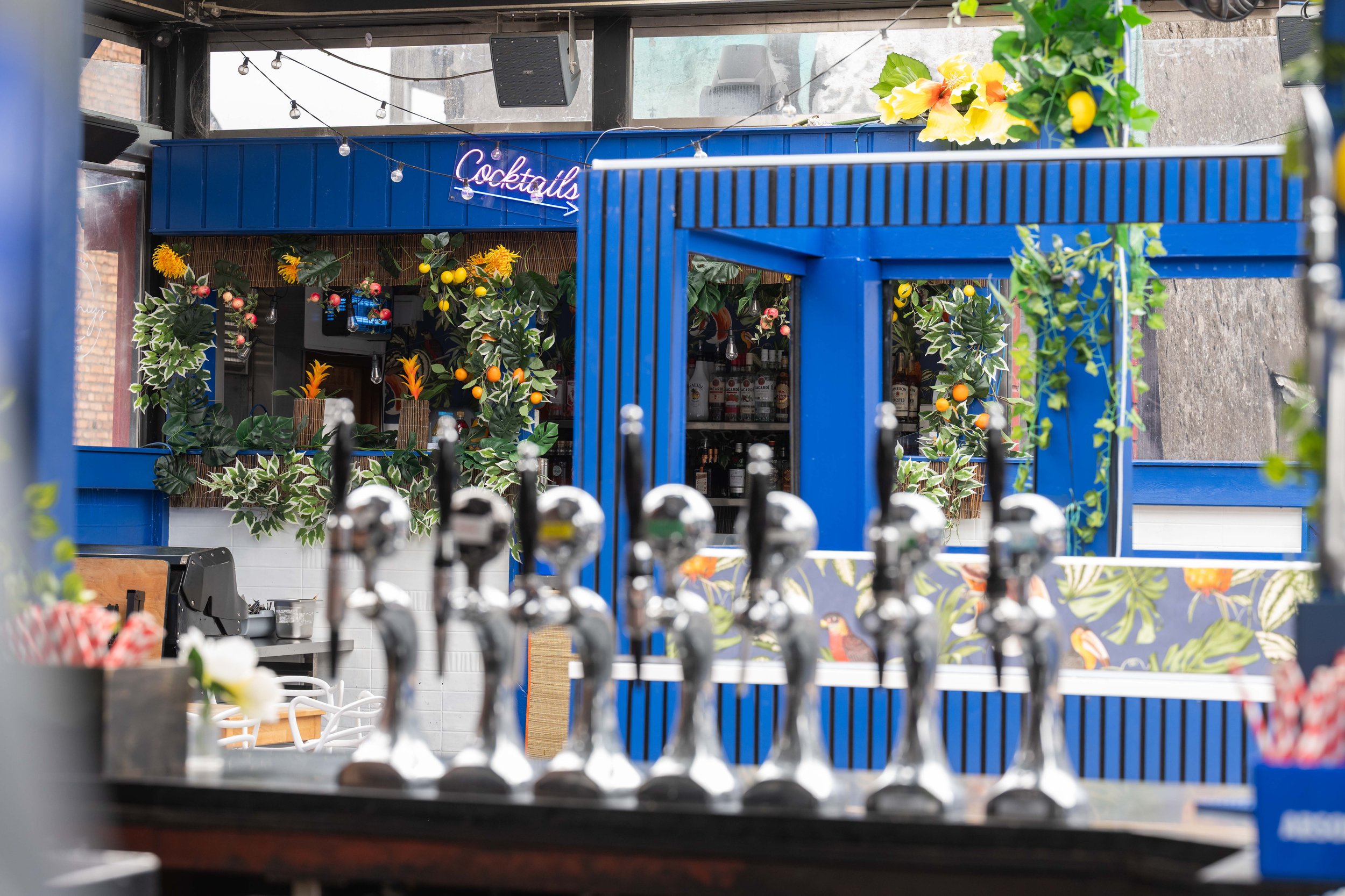 View of a bar inside a restaurant with a neon sign that reads "Cocktails", decorated with artificial plants and flowers, including orange and yellow blossoms, with beer taps in the foreground.