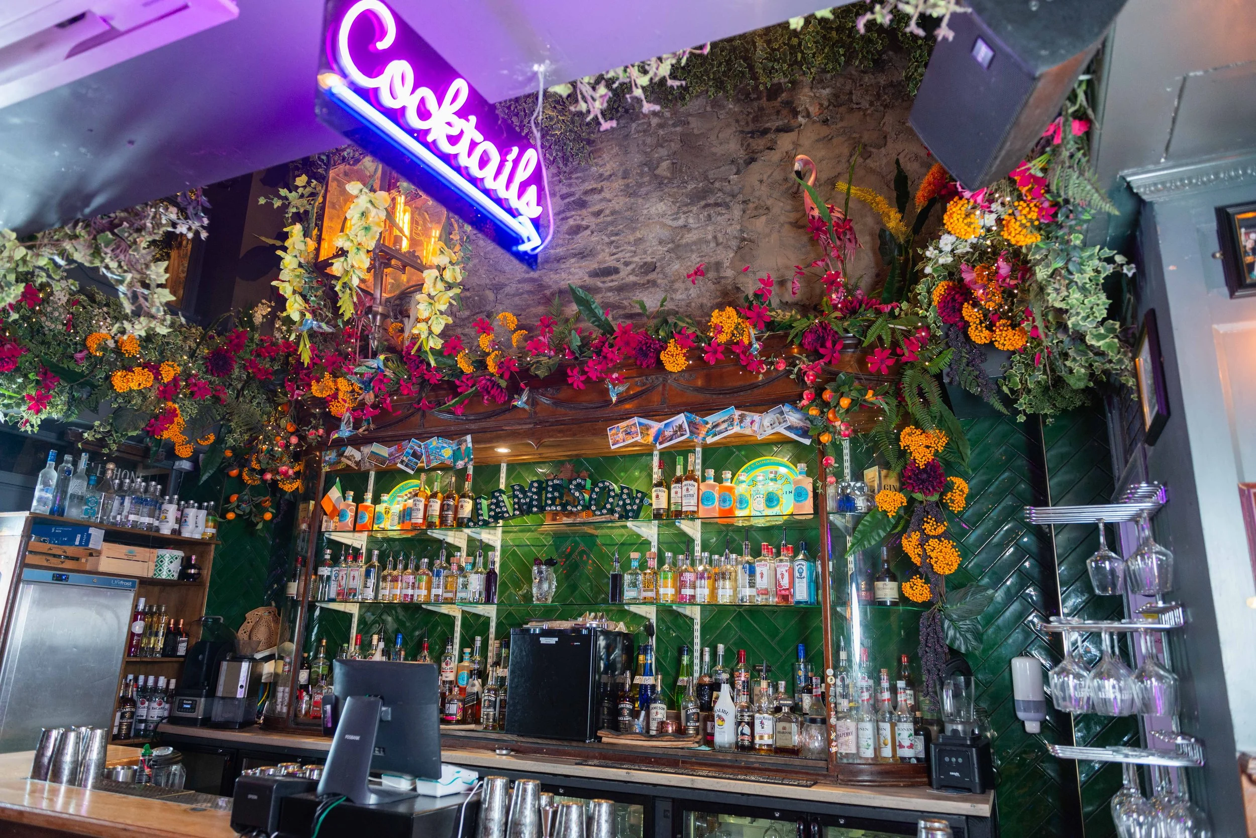 A colourful bar with shelves of liquor bottles, decorated with vibrant flowers and plants, and a neon sign that says 'Cocktails' hanging above.