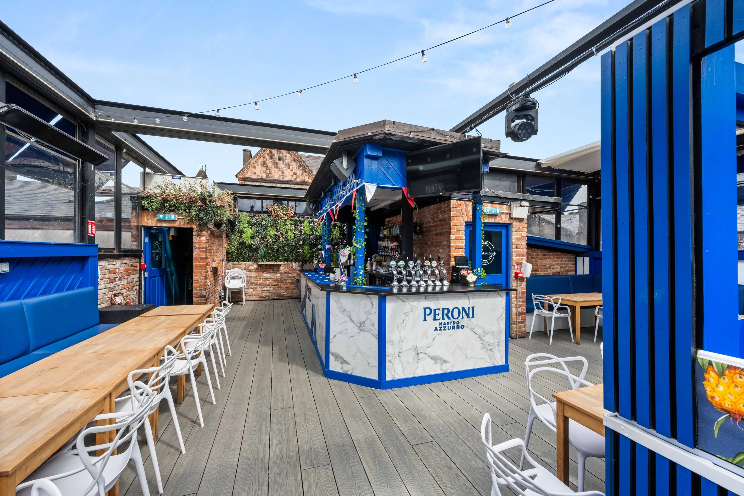 An outdoor rooftop bar with a blue and white theme, featuring a marble front bar, beer taps, and surrounding tables with white chairs and blue seating. String lights hang above, and greenery decorates the brick walls.