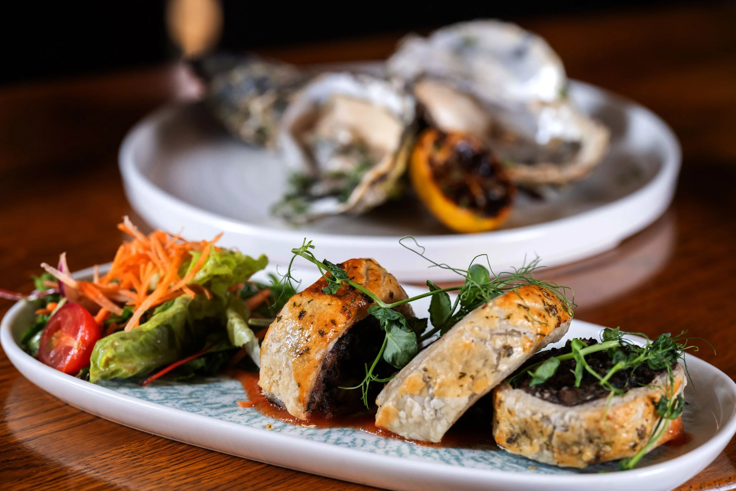 Plate of stuffed chicken breasts garnished with microgreens, served with a side salad of lettuce, cherry tomatoes, and shredded carrots, with oysters and lemon in the background.