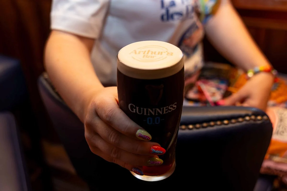 Person holding a pint glass of Guinness beer with an Arthur's Day logo on the foam. The person has colorful nail polish and a multicolored bracelet, sitting in a cozy, dimly lit setting.