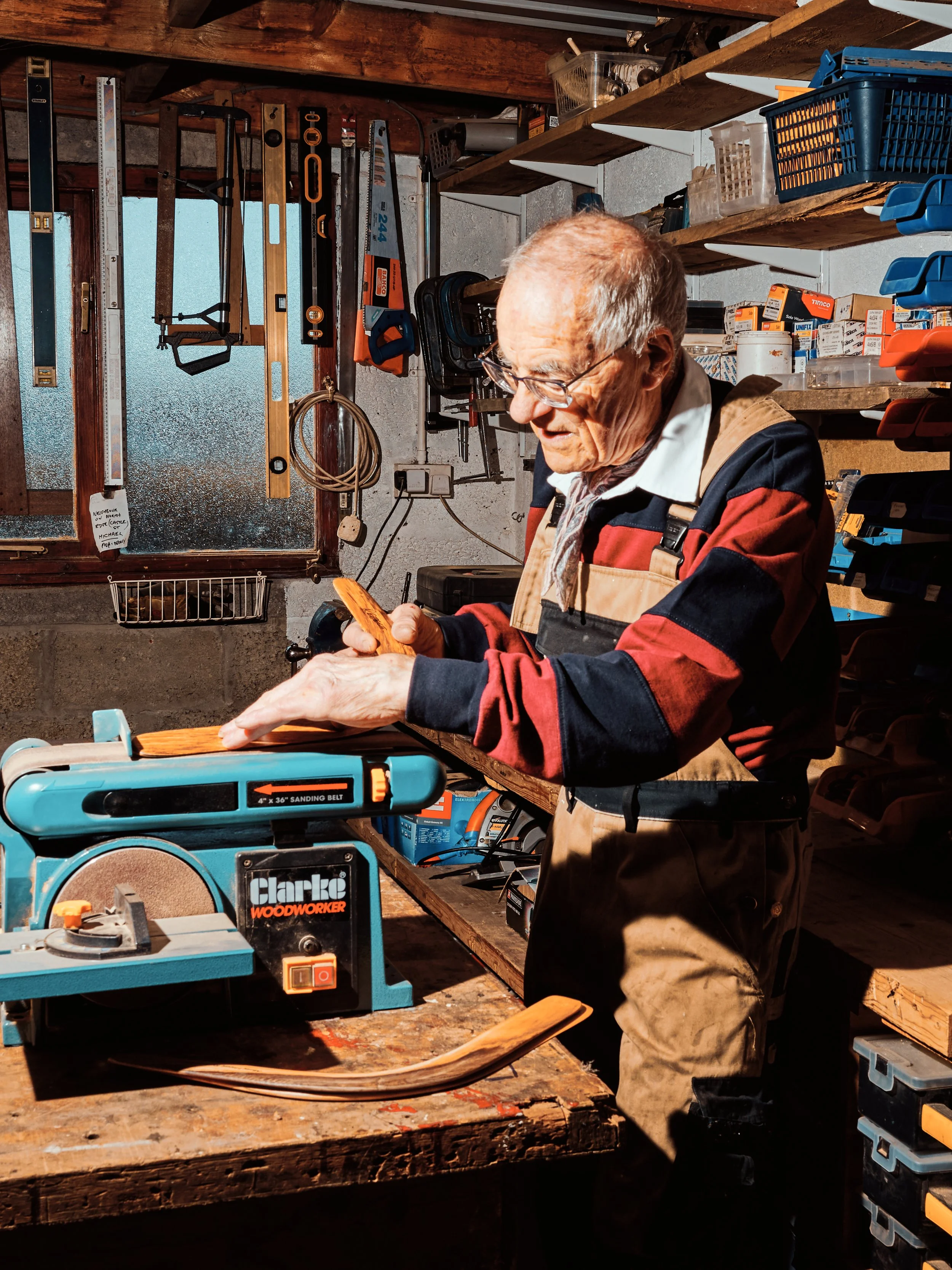 An elderly man working in a cluttered woodworking workshop, surrounded by tools, shelves, and equipment, as he examines a piece of wood.
