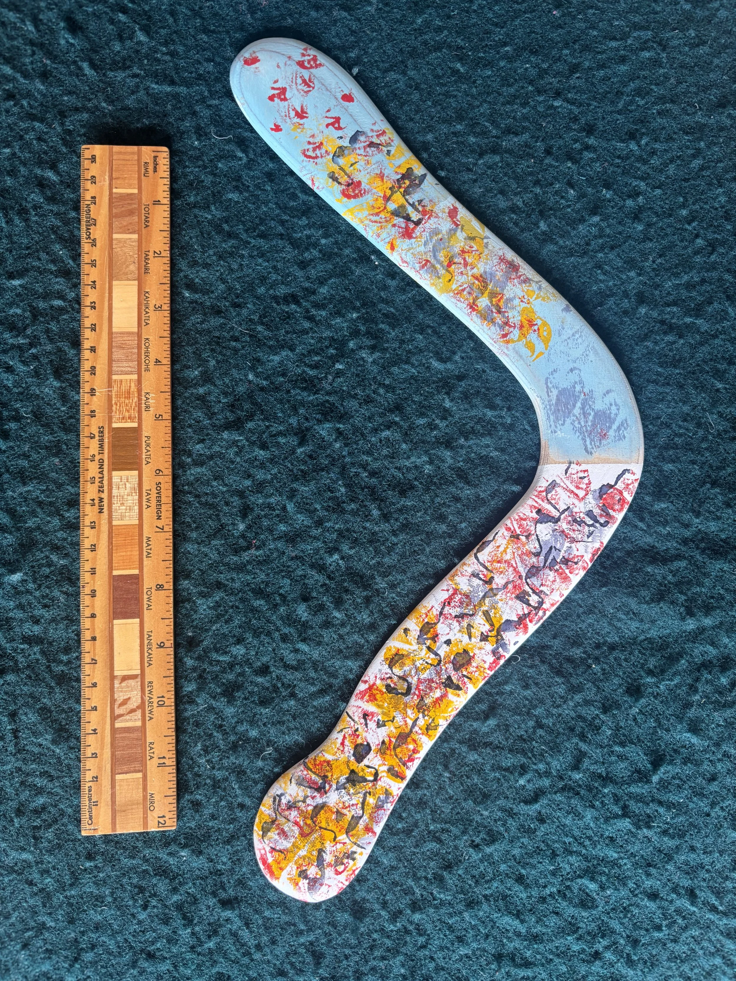 A painted, curved object resembling the letter S on a dark textured surface, with a wooden ruler placed beside it for scale.