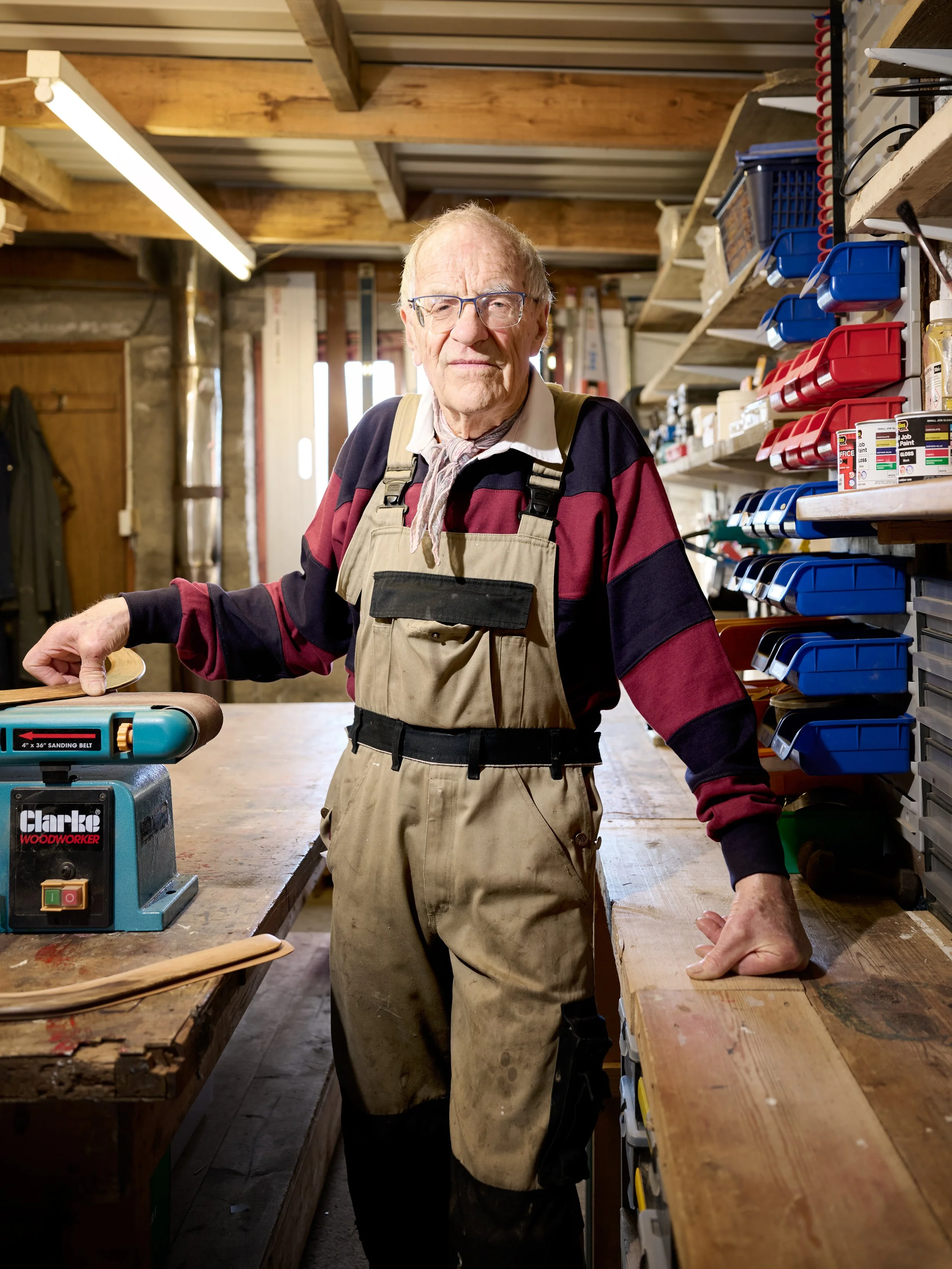 An elderly man with glasses and a beige apron working in a woodworking shop, standing at a workbench with tools and storage shelves around him.