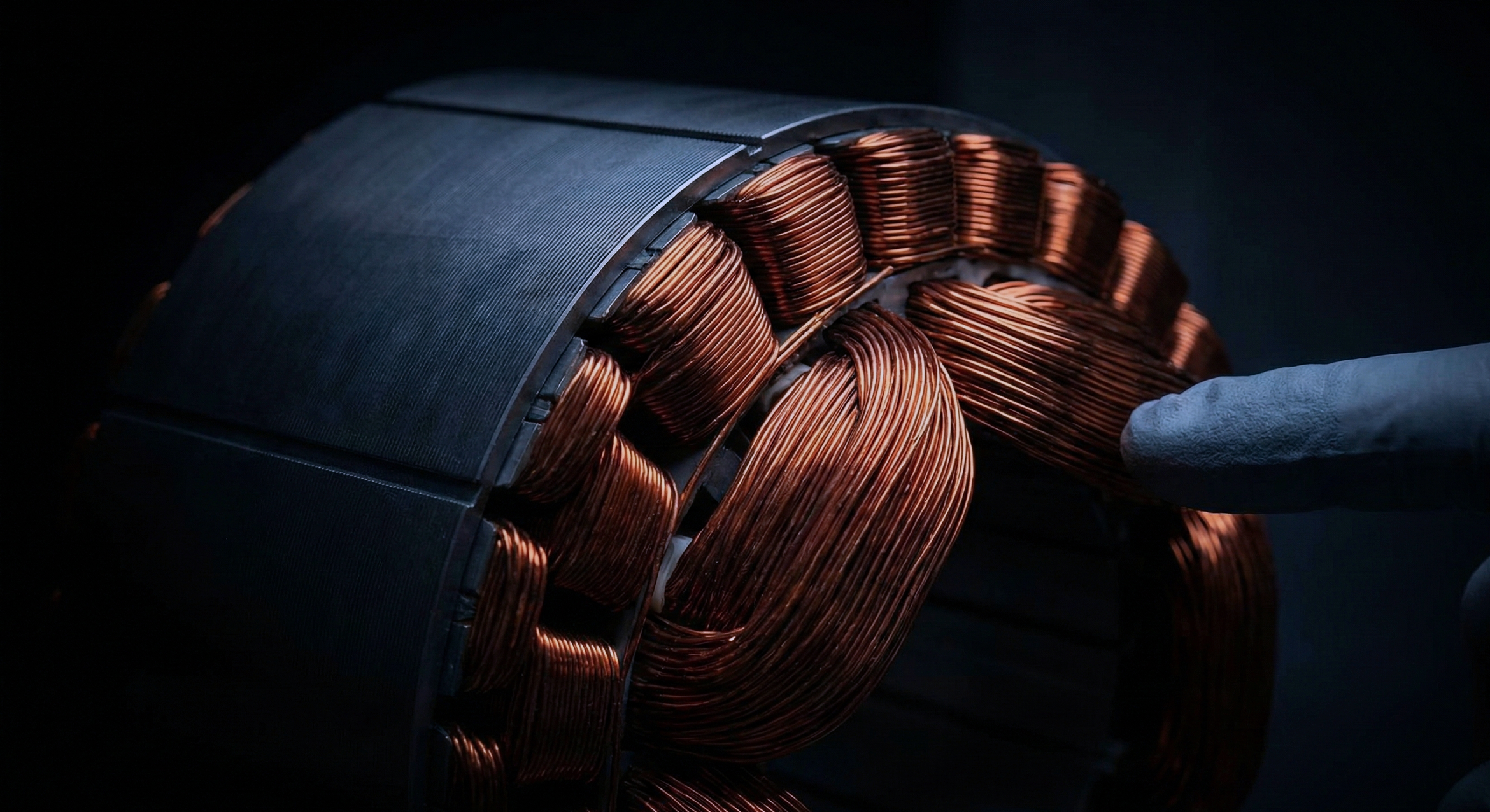 Close-up of a large electrical motor coil with copper wire windings and a gloved hand touching it.