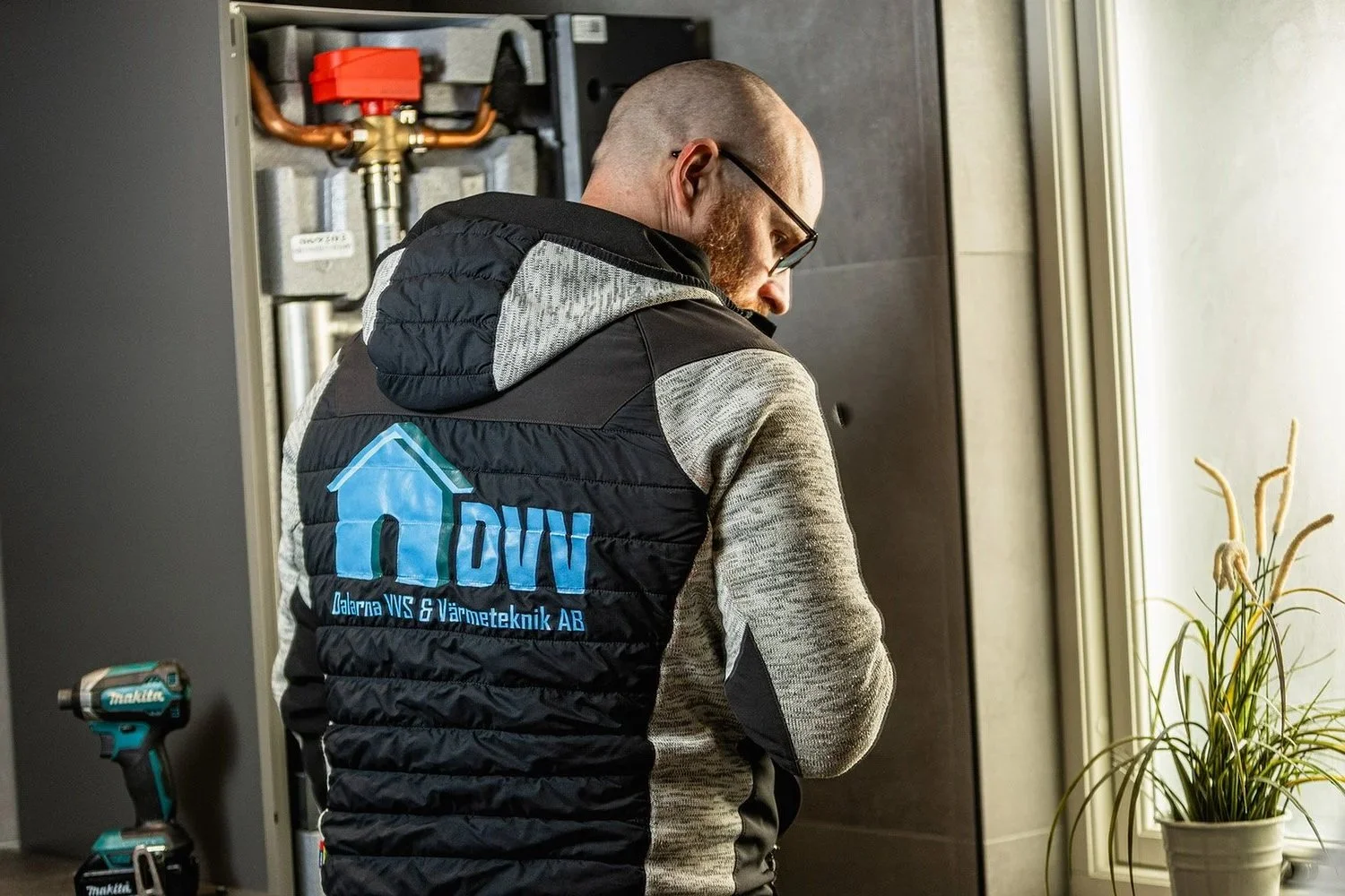 En man i svart och grå jacka, med glasögon, arbetar på en apparat eller maskin, troligtvis inom värmeteknik, inomhus nära ett fönster med en växt på fönsterbrädan.