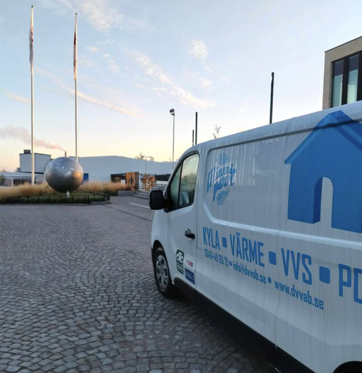 En vit servicebil med text om kyla, värme, VVS och andra tjänster parkerad på en stenlagd yta utanför en modern byggnad. I bakgrunden syns en staty av en jordglob med tre flaggstänger.