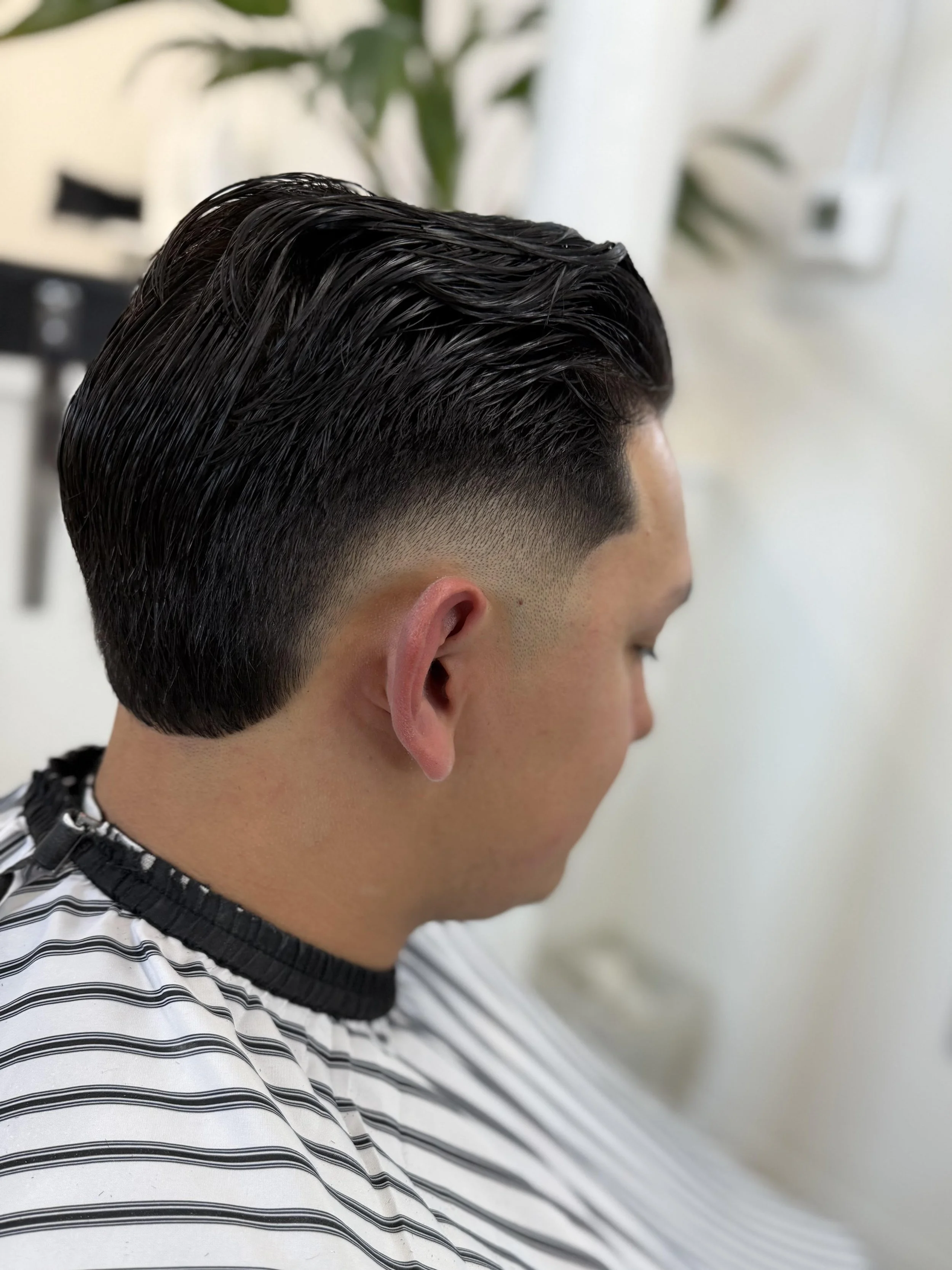 Side view of a man with a freshly styled, slicked-back haircut with faded sides, wearing a striped barber cape in a barber shop.
