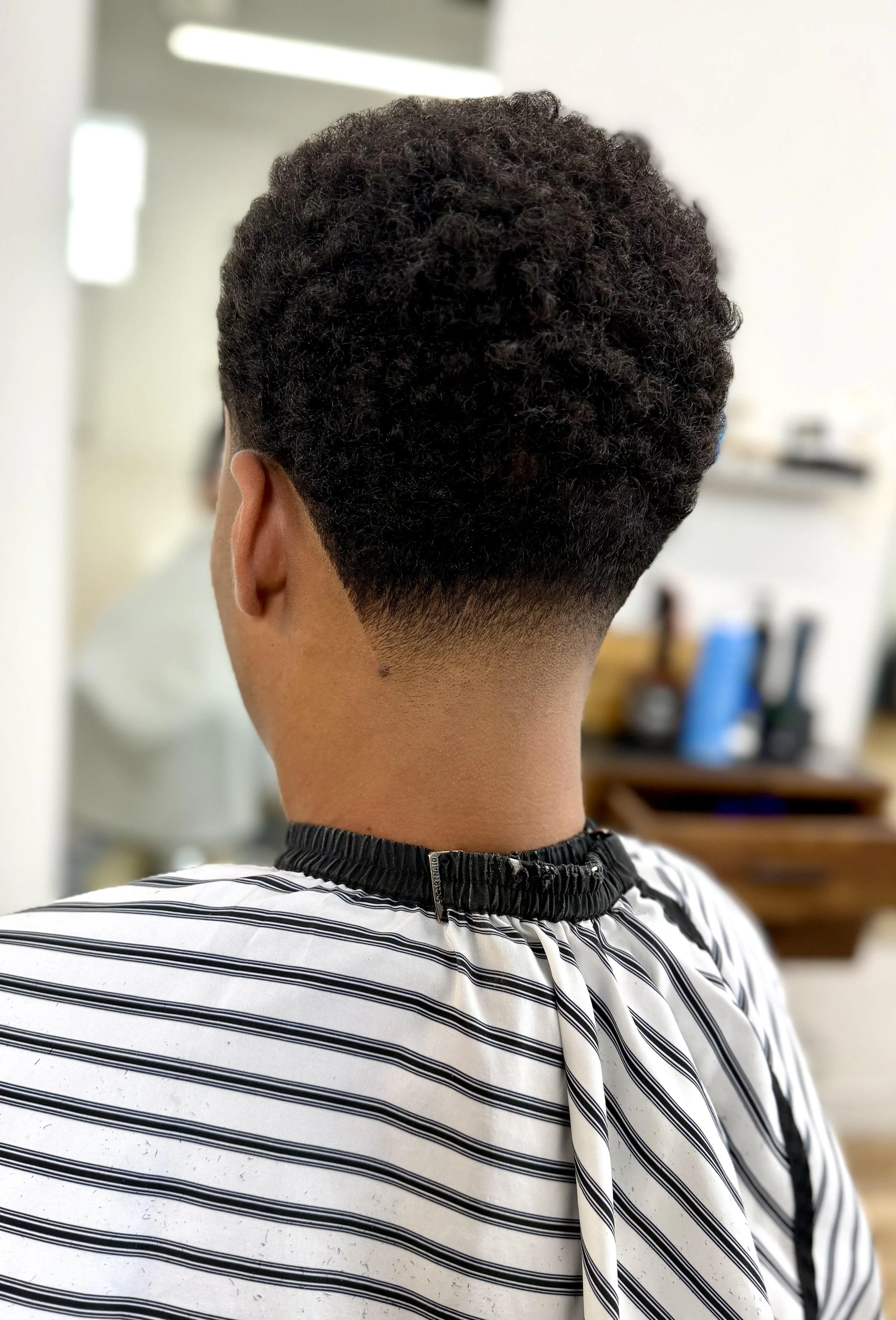 Back of a person's head showcasing a fresh haircut with a fade and curly hair texture, wearing a barber cape with black and white stripes in a barber shop.