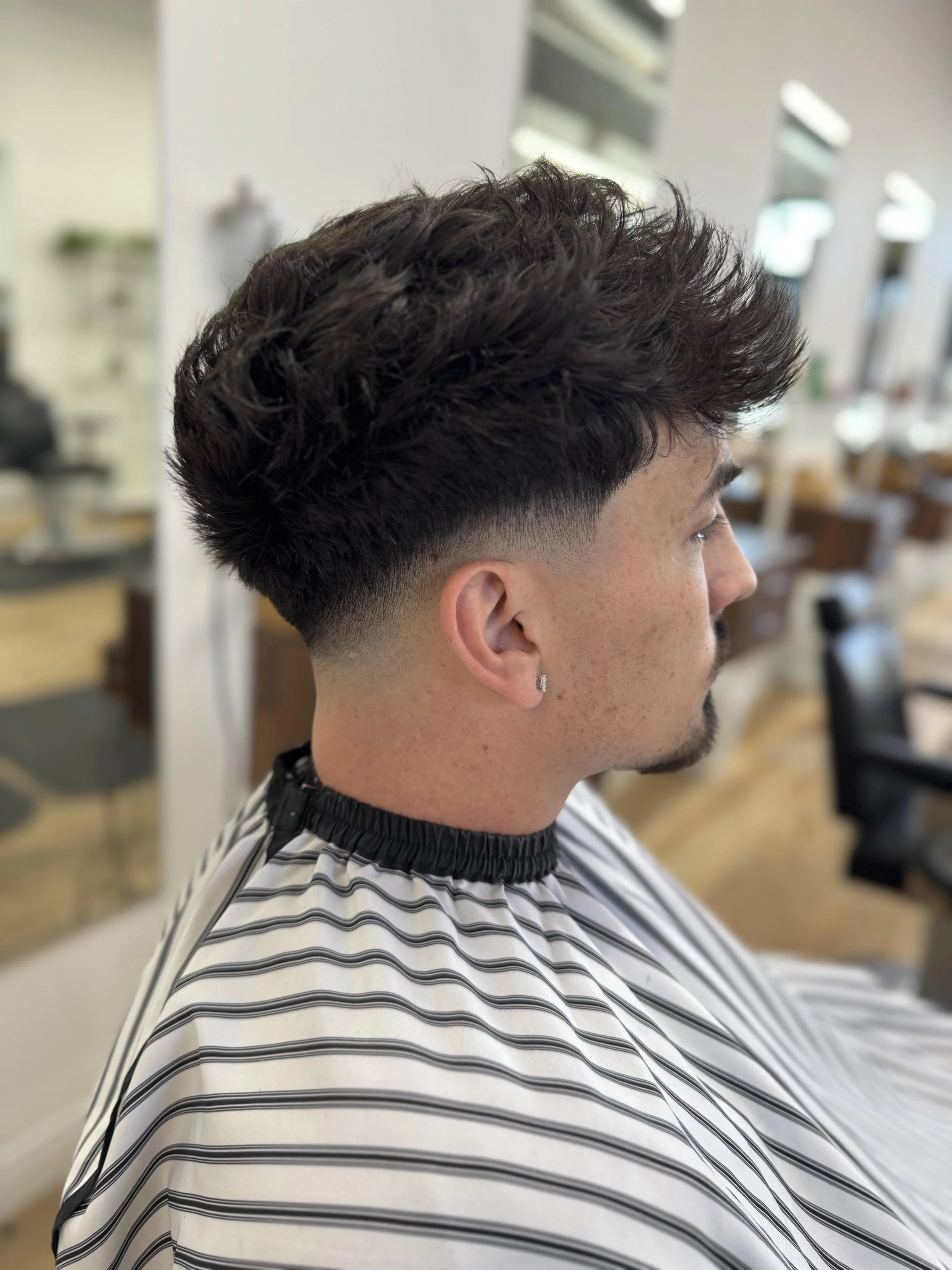 Side profile of a young man with a textured curly hairstyle and a faded undercut, seated in a hair salon.