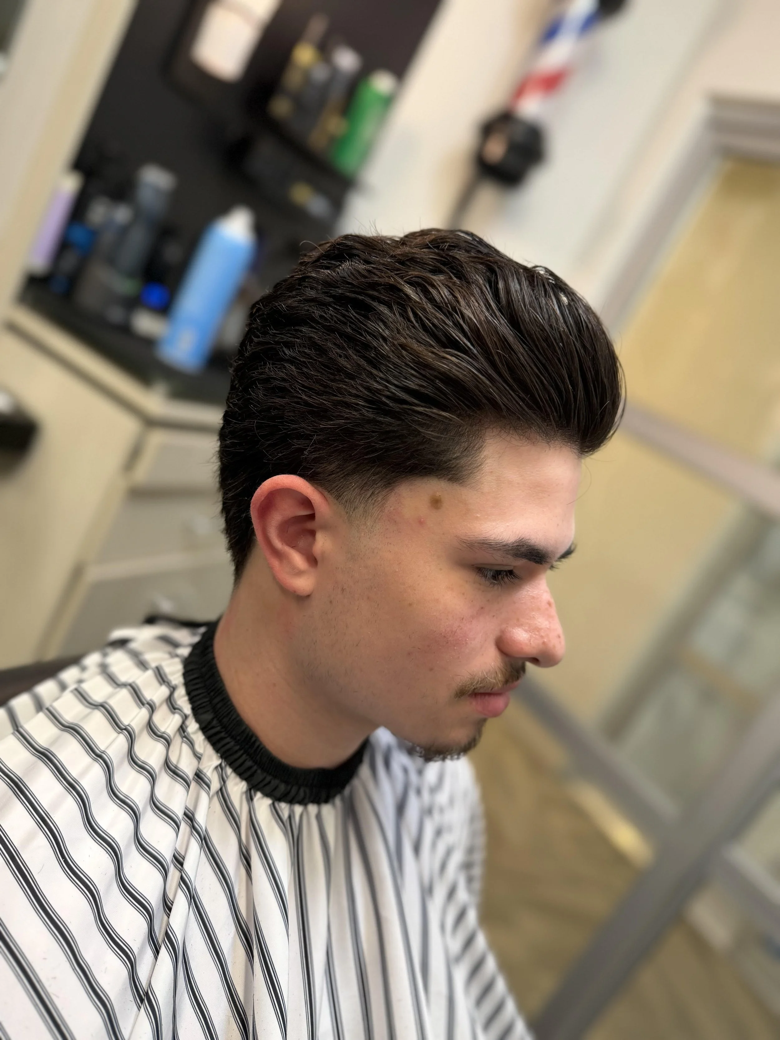 A young man sitting in a barber's chair, with freshly styled dark brown hair, wearing a black and white striped barber cape, in a barbershop.
