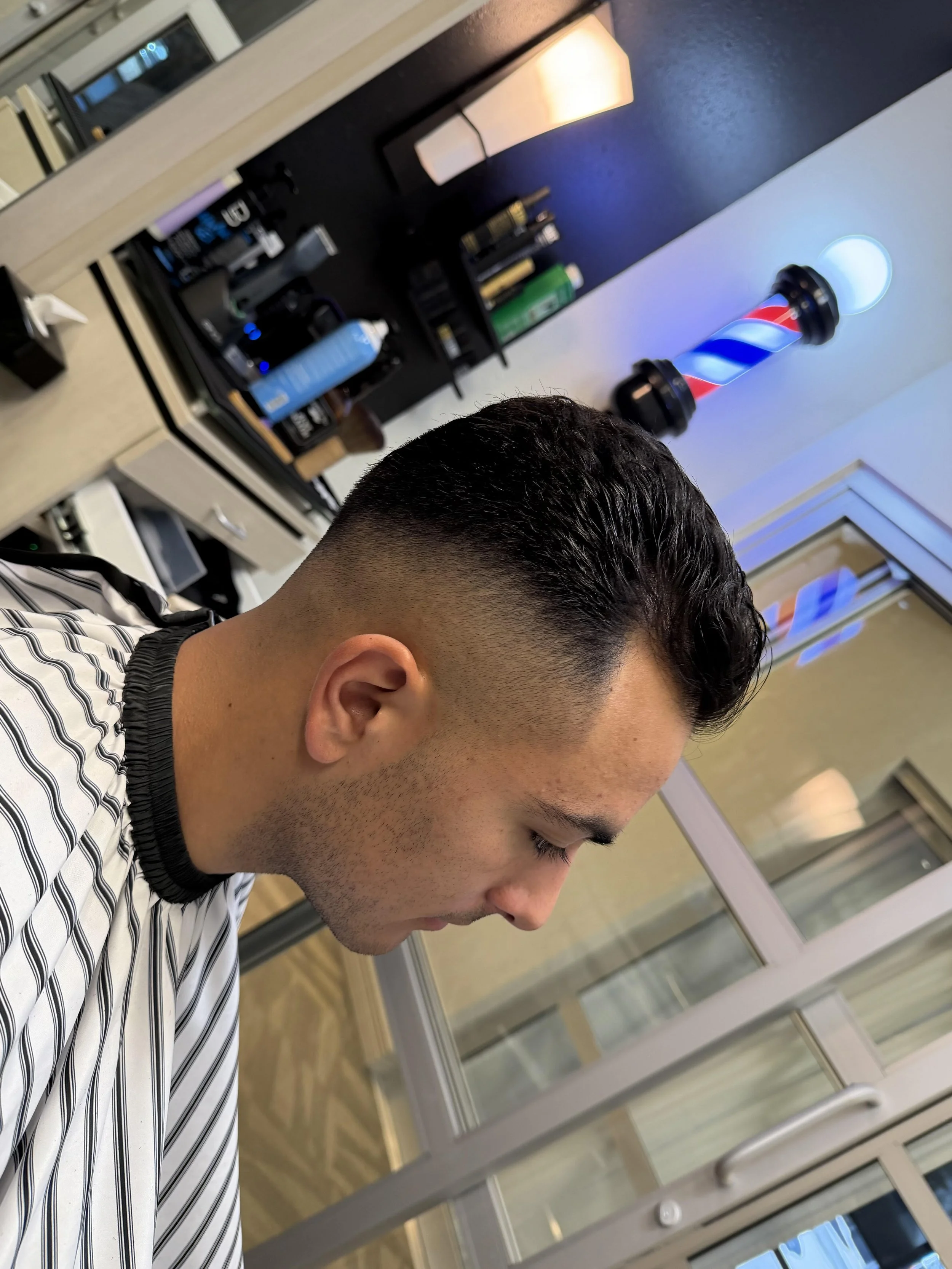 Man sitting in barber shop chair with freshly cut hairstyle, mirror and barber tools visible on counter, barber pole outside window.