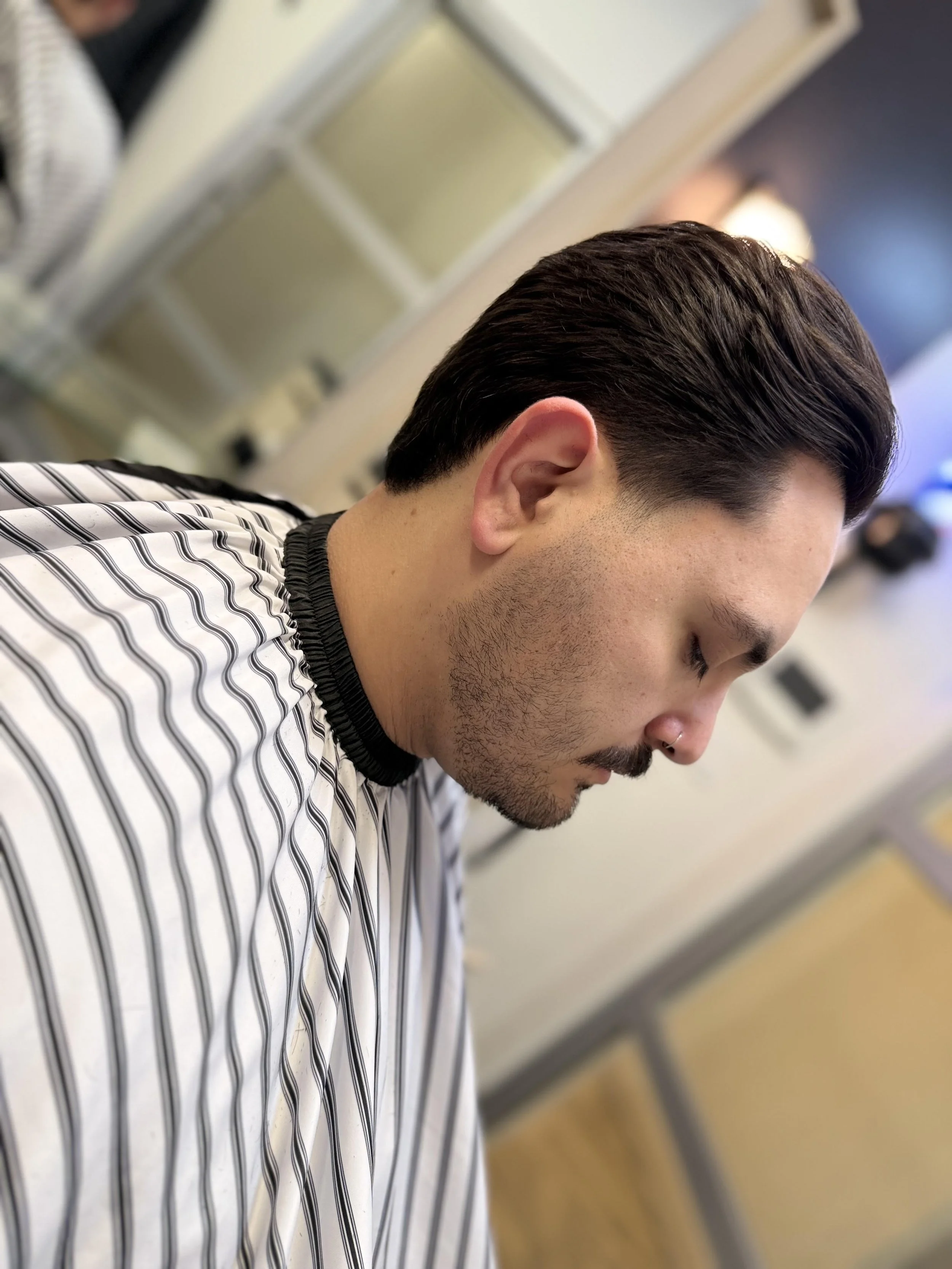 Side profile of a man with combed dark hair and a beard, sitting in a barbershop with a striped barber cape.