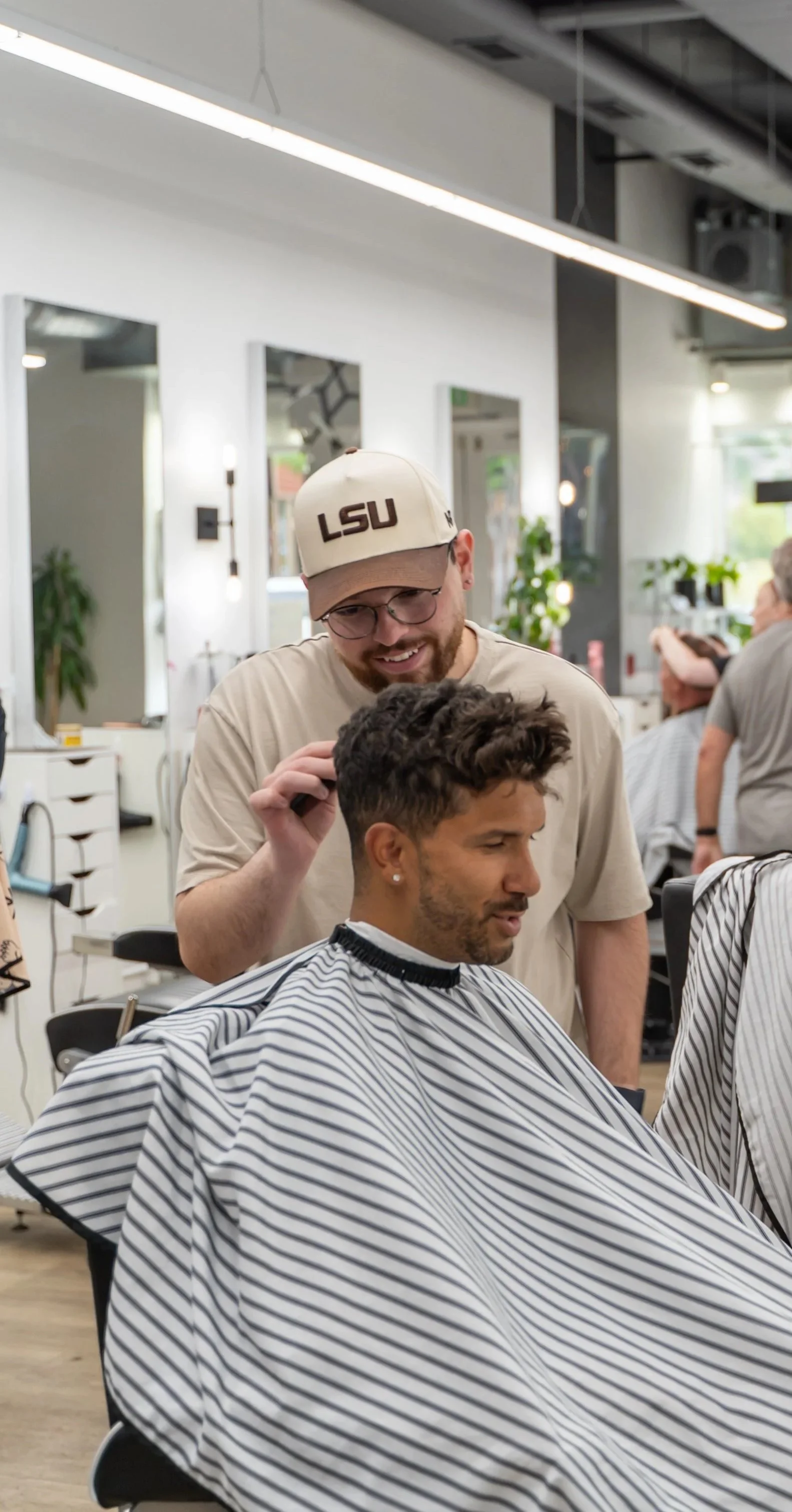 A barber styles a customer's hair in a modern barbershop. The customer is seated with a striped cape, and the barber is smiling while working.