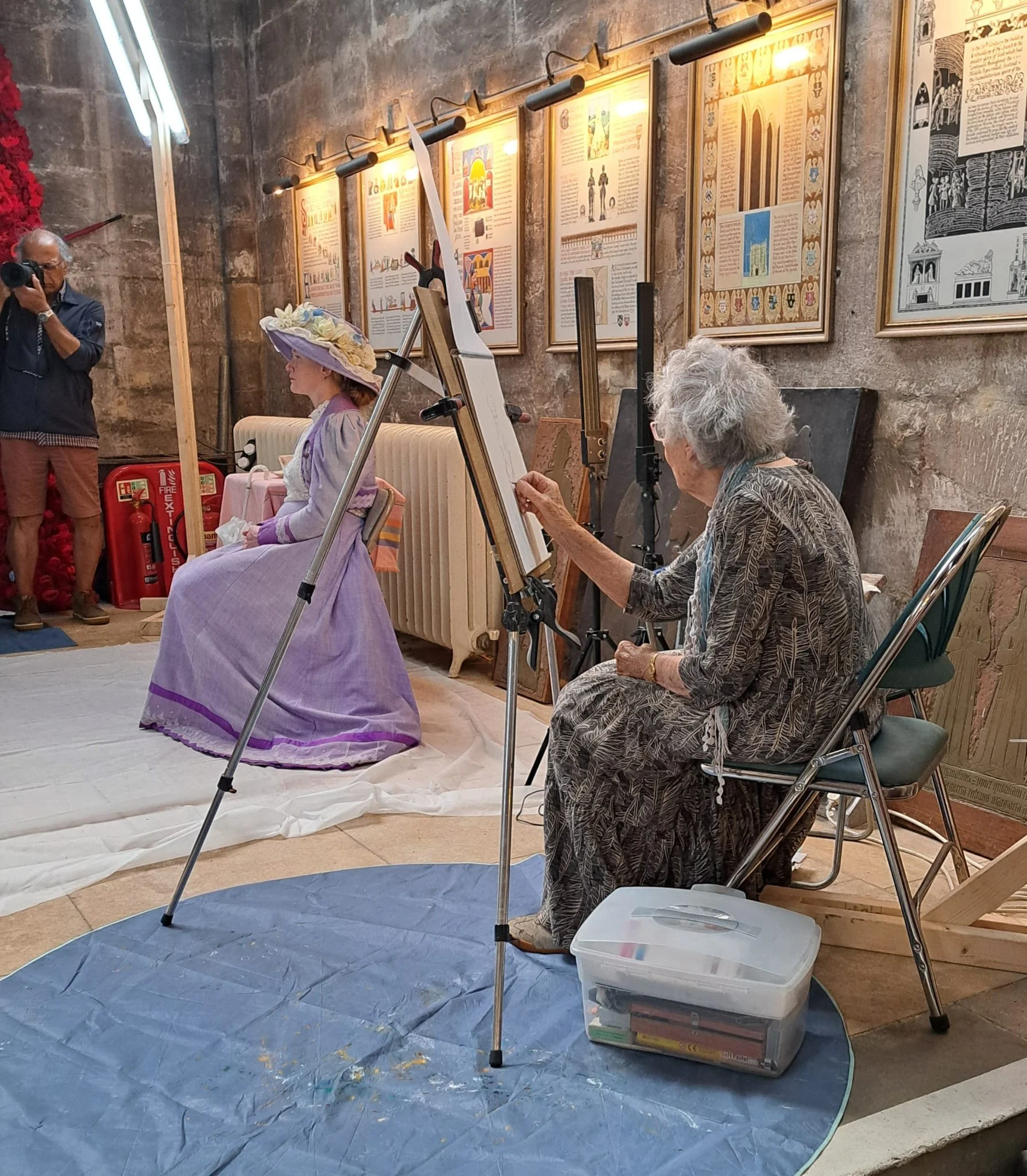 An elderly woman painting a portrait of a woman dressed in a vintage purple gown and large hat, seated on a bench, in an art studio setting with framed artwork on the wall. A man is taking a photograph of the scene.