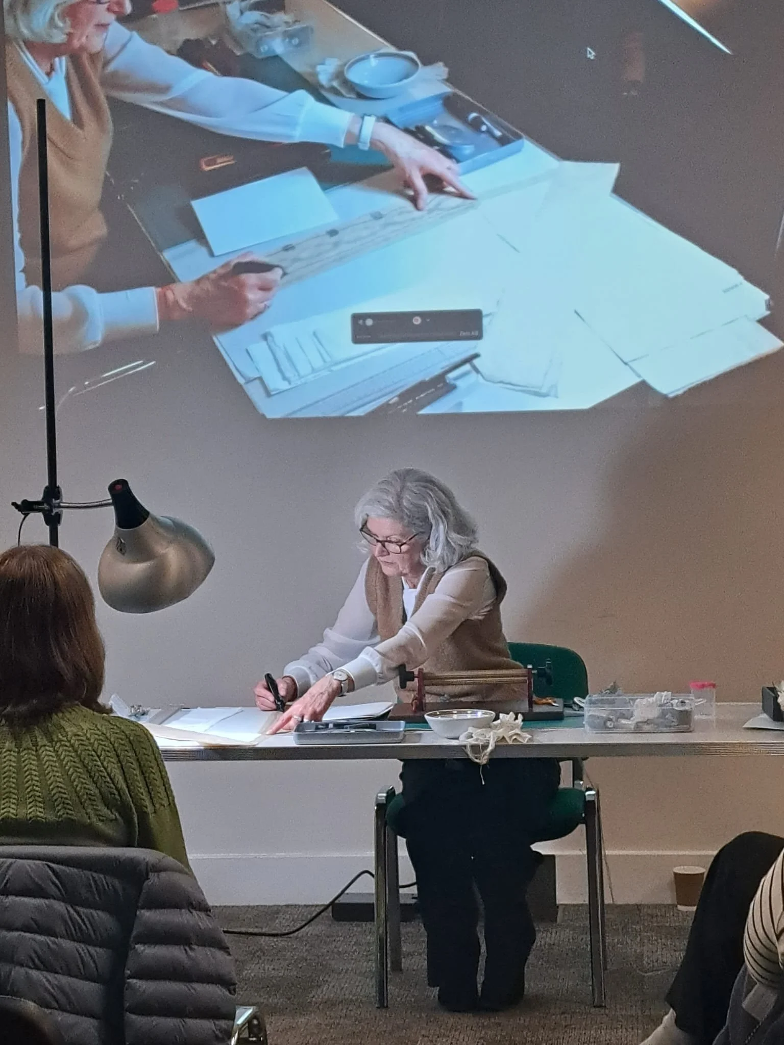 A woman with gray hair and glasses sitting at a table, writing or drawing on paper. There is a large projection on the wall behind her showing her working at the table. Several people are seated in front of her, and the table has various tools and ma