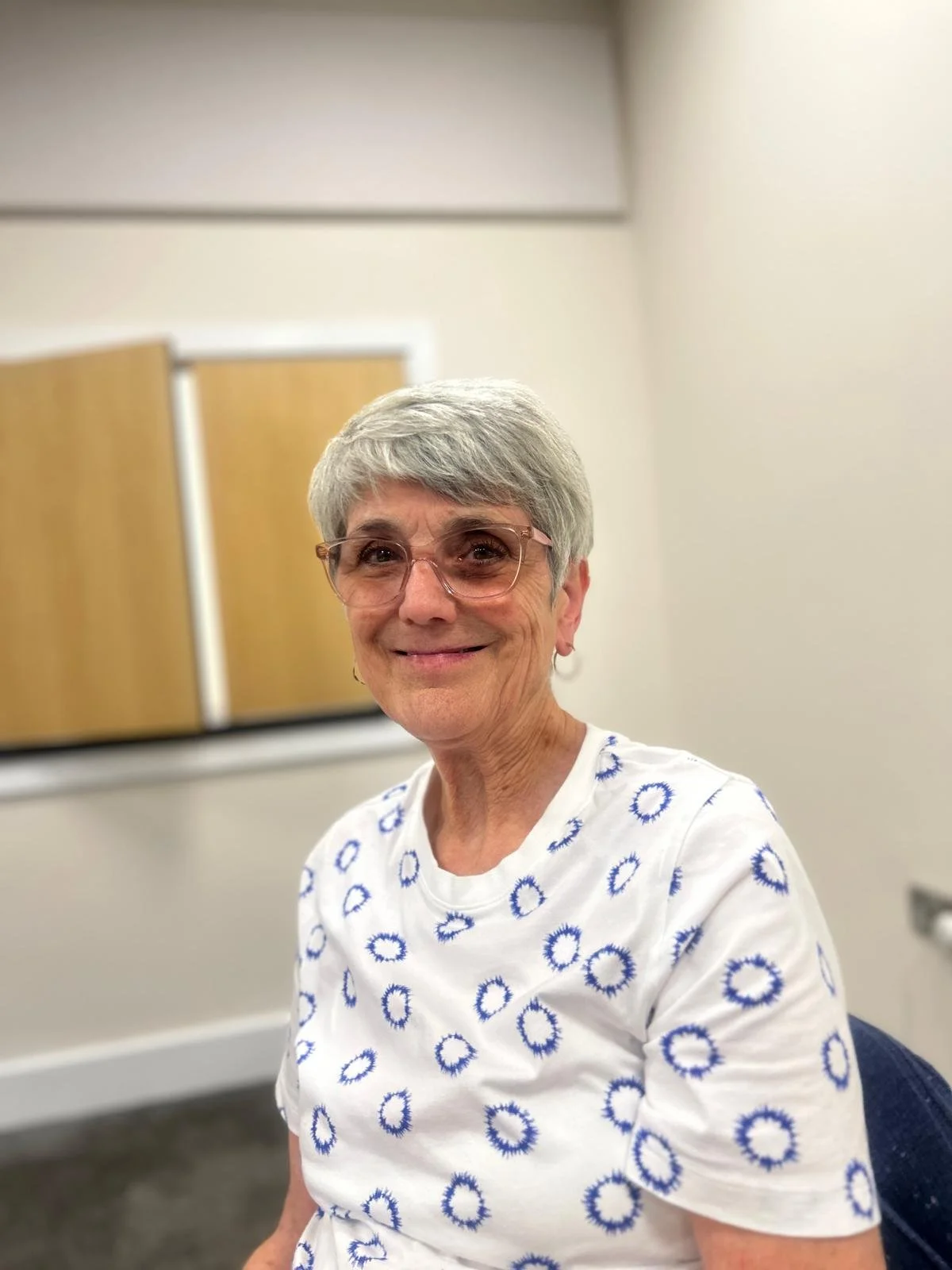 An older woman with short gray hair, wearing glass glasses and a white shirt with blue circular patterns, smiling at the camera.