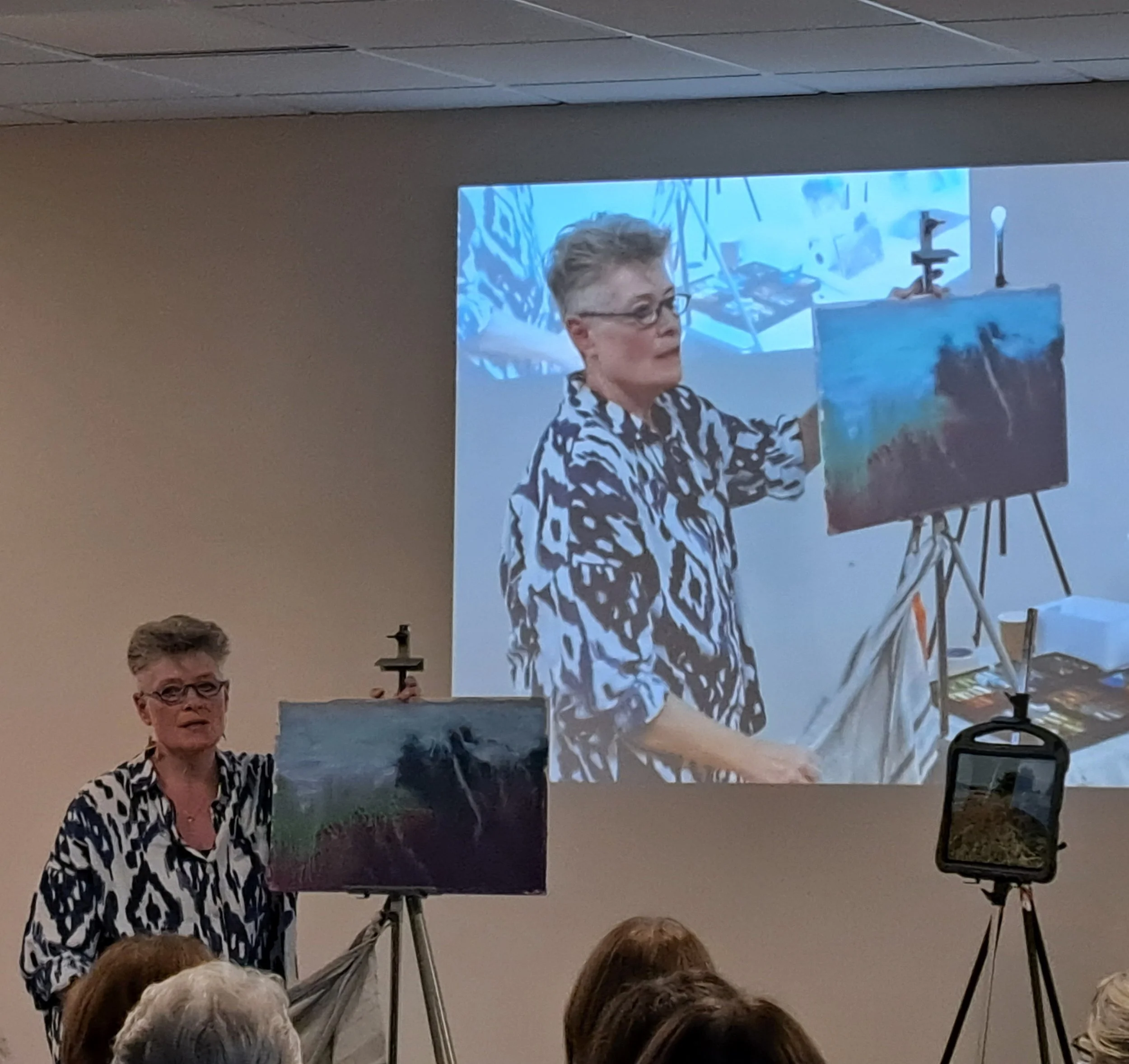 An older woman with short hair and glasses standing beside an easel with a partially finished landscape painting, and a projected image of her working on the painting on a large screen behind her.