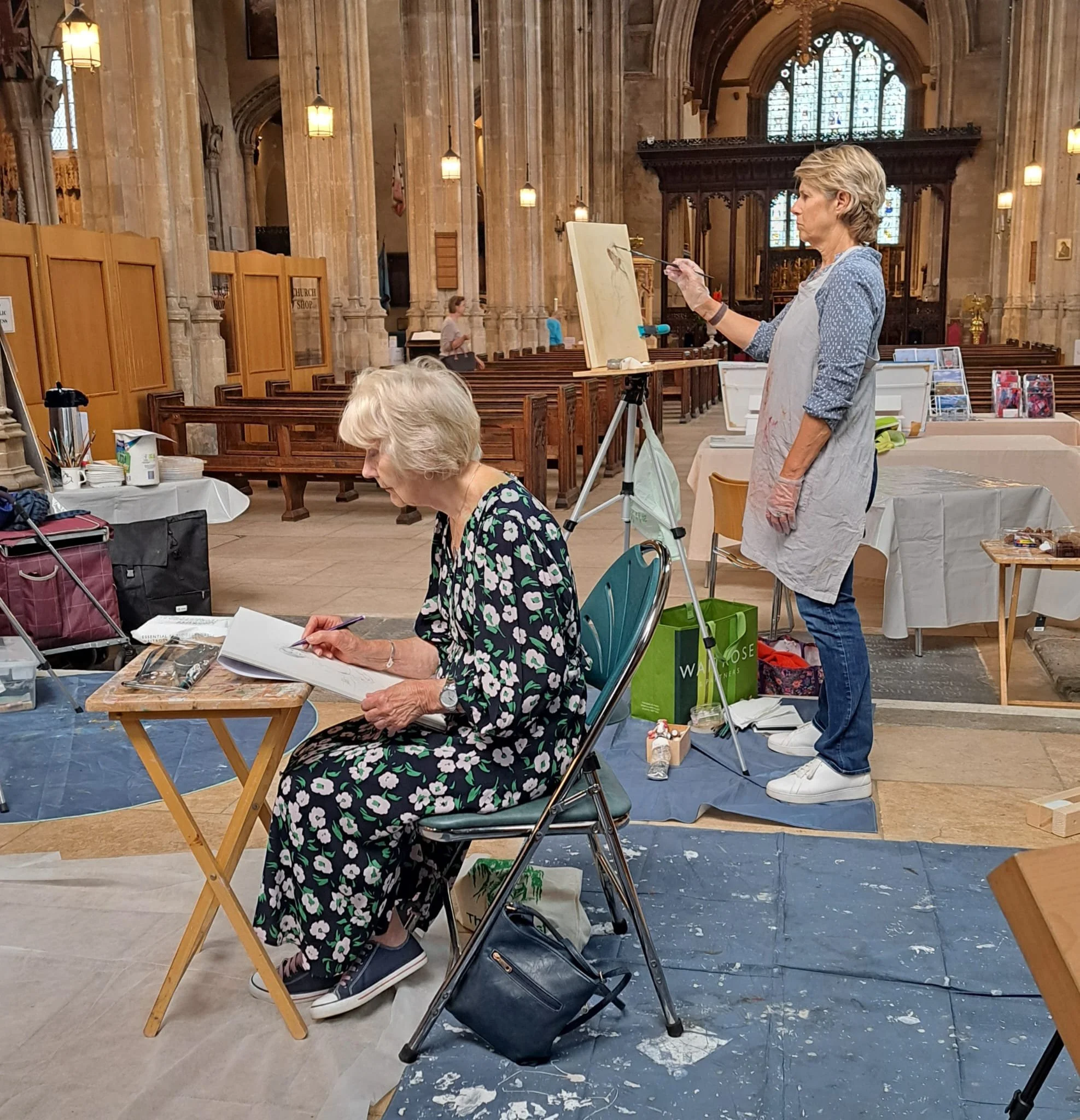 An elderly woman in a floral dress drawing in a sketchbook, with a woman in a grey apron painting on an easel in a church interior.