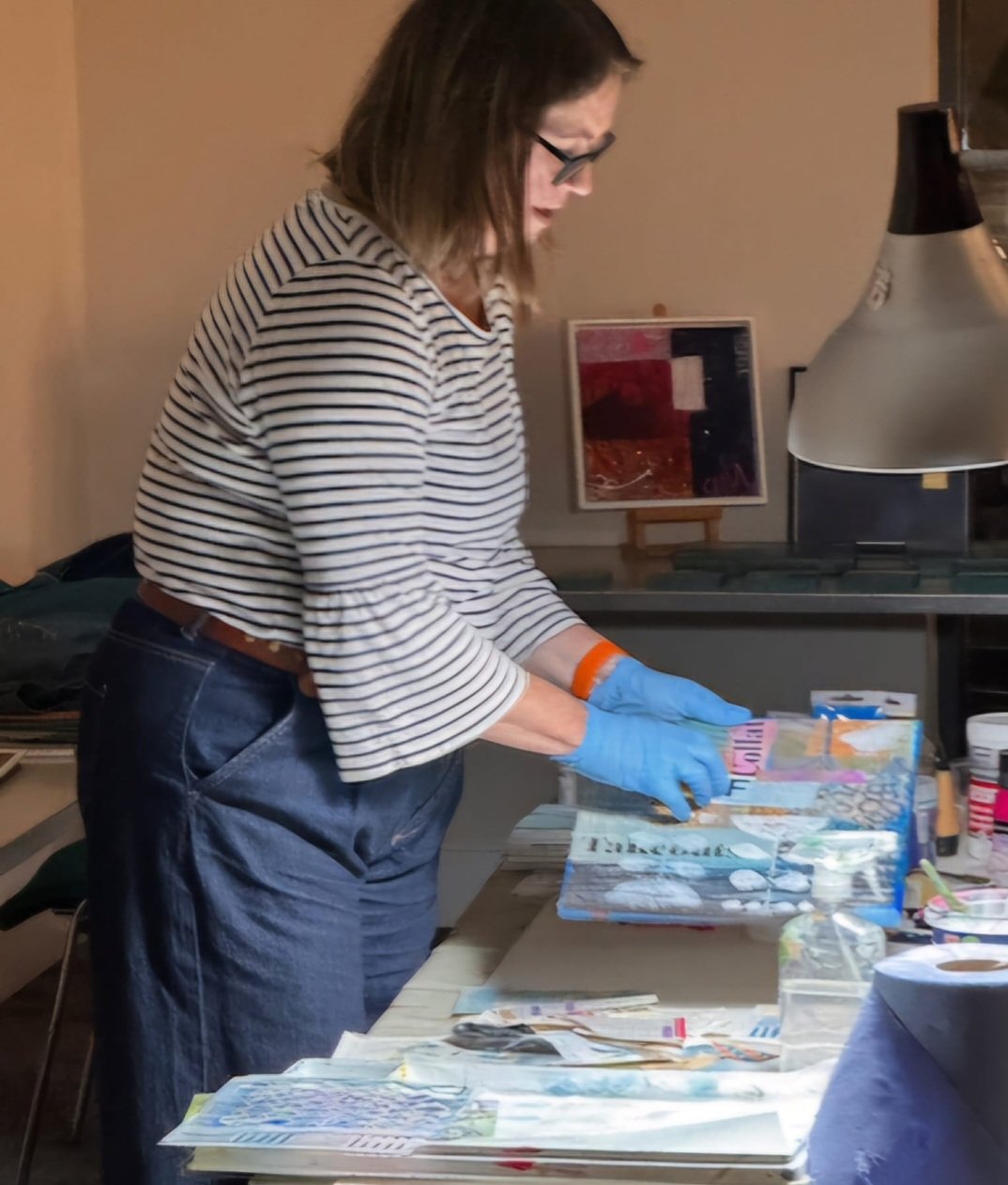 A woman wearing a striped shirt and glasses is working with colorful paper or artwork on a table, wearing blue gloves.
