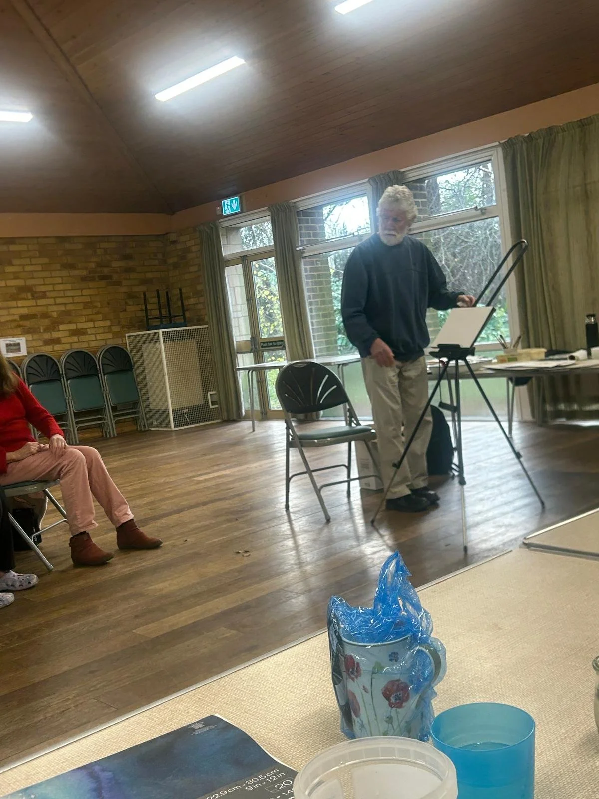 An elderly man with glasses and white hair, standing and writing on a whiteboard or flip chart in a room with large windows and wooden floors, while seated people observe.