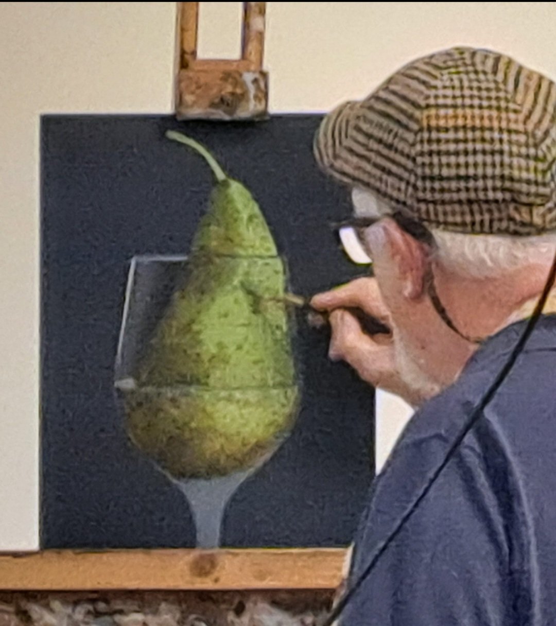 A man wearing a hat and glasses working on an art piece featuring a large pear. The pear is cut in half and displayed with a black background, mounted on a vertical surface.