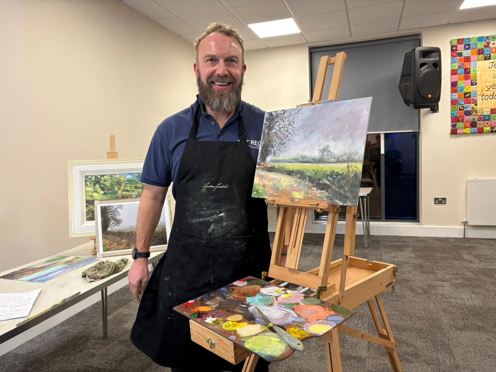 A man with a beard and short light brown hair, wearing a black apron and a dark blue polo shirt, is standing in a room with beige walls and gray carpet. He is smiling and holding a palette with various paint colors. In front of him is an easel with a