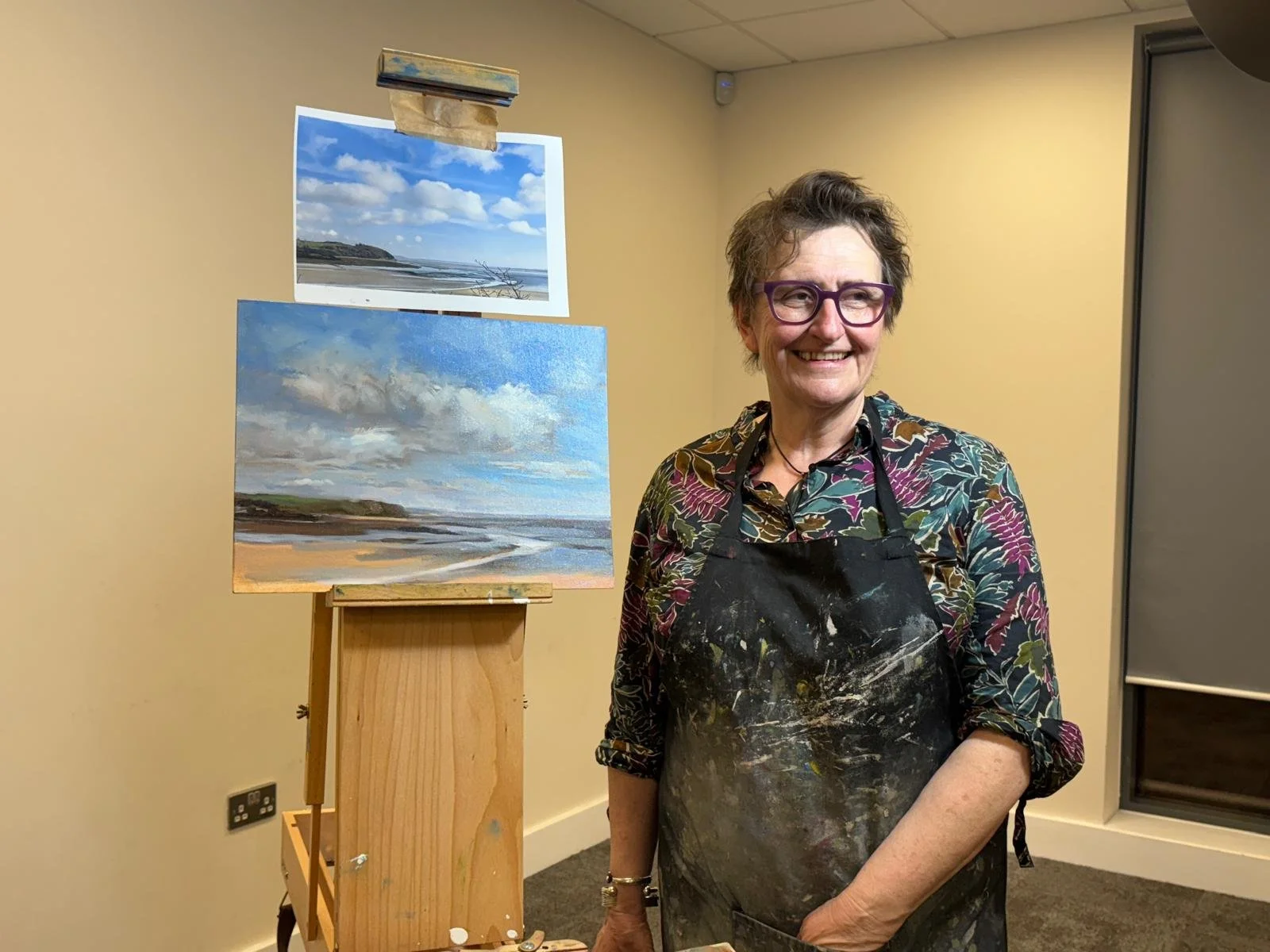 A woman with short hair, purple glasses, and a floral shirt smiling in front of two landscape paintings, one on an easel and one mounted on the wall, in an indoor art studio or gallery.