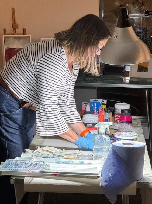 A woman wearing a striped long-sleeve shirt and gloves is working at a table with various art supplies, possibly creating a mixed media artwork, in a well-lit room.