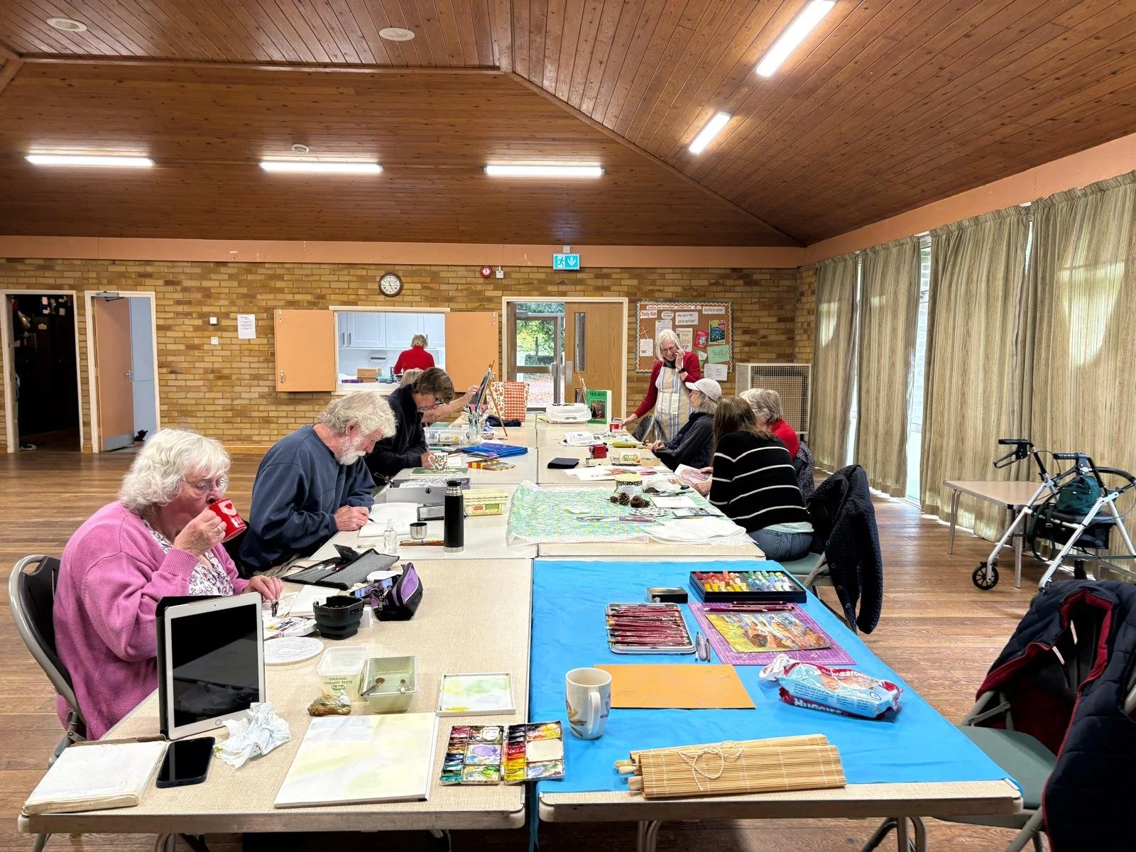 A group of people sitting at a long table in a community room, engaged in arts and crafts activities. There are art supplies, a laptop, and personal items on the table. A woman standing at the far end of the table is wearing an apron, and a walker is visible near the window on the right side.