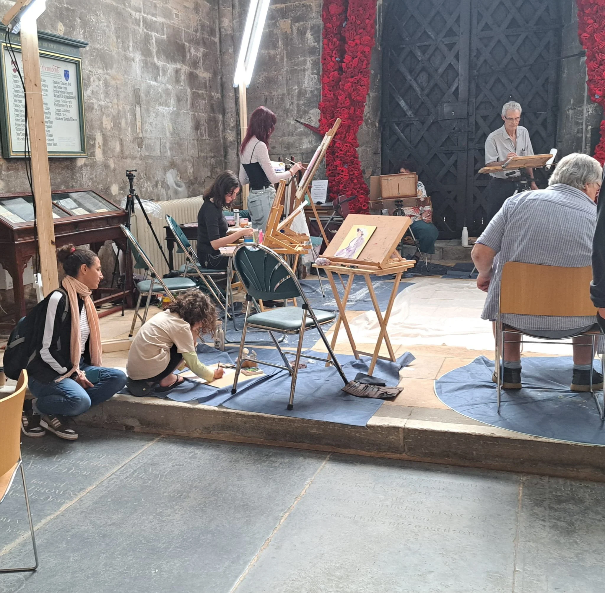 Artists painting and drawing on easels and tables in a gallery or museum setting with red floral decorations and stone walls.
