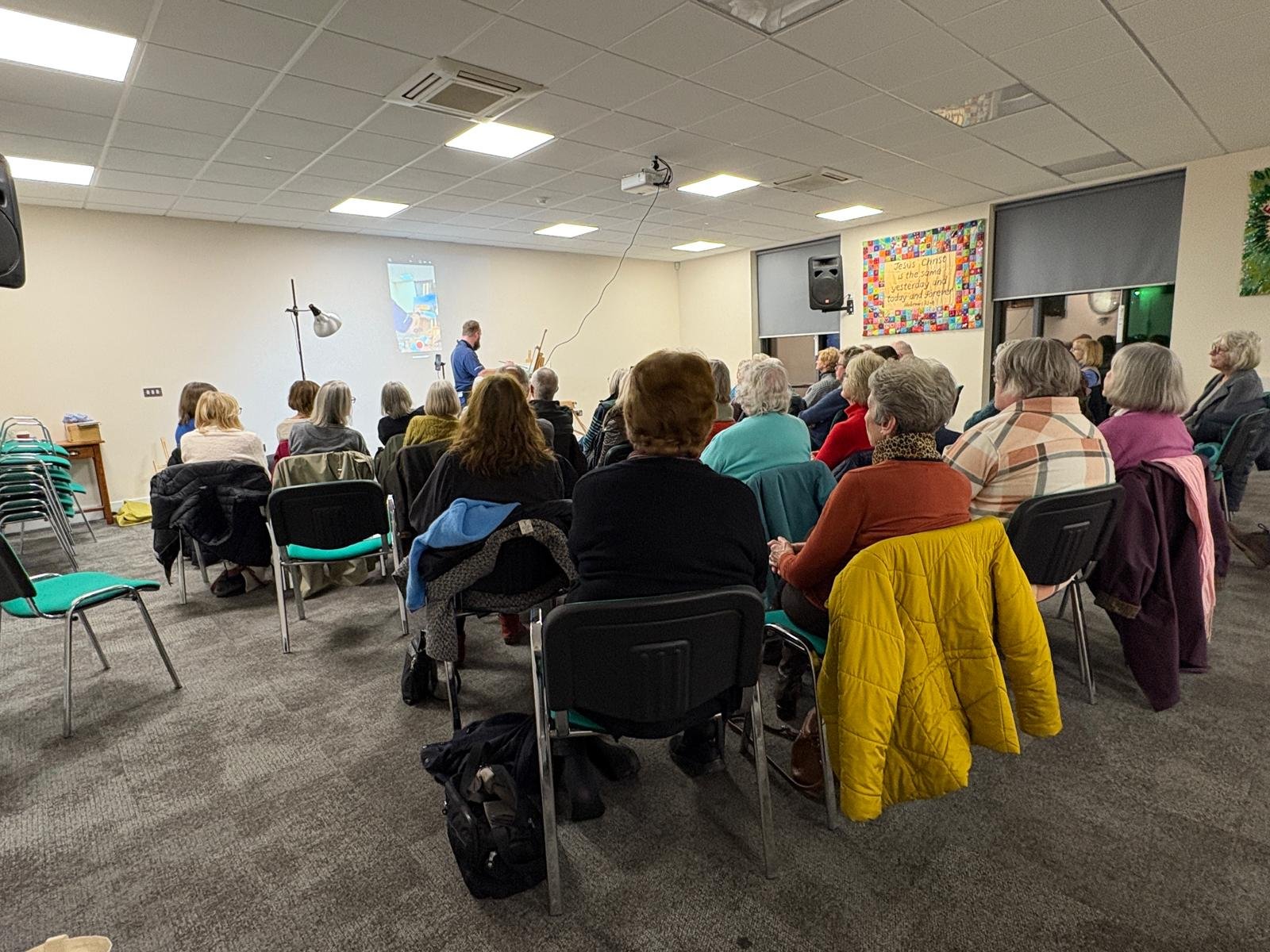 Group of people seated in a conference room, watching a person playing the piano at the front. The room has a projector screen with a colorful image projected on the wall, and colorful decorations, including a Christmas banner.