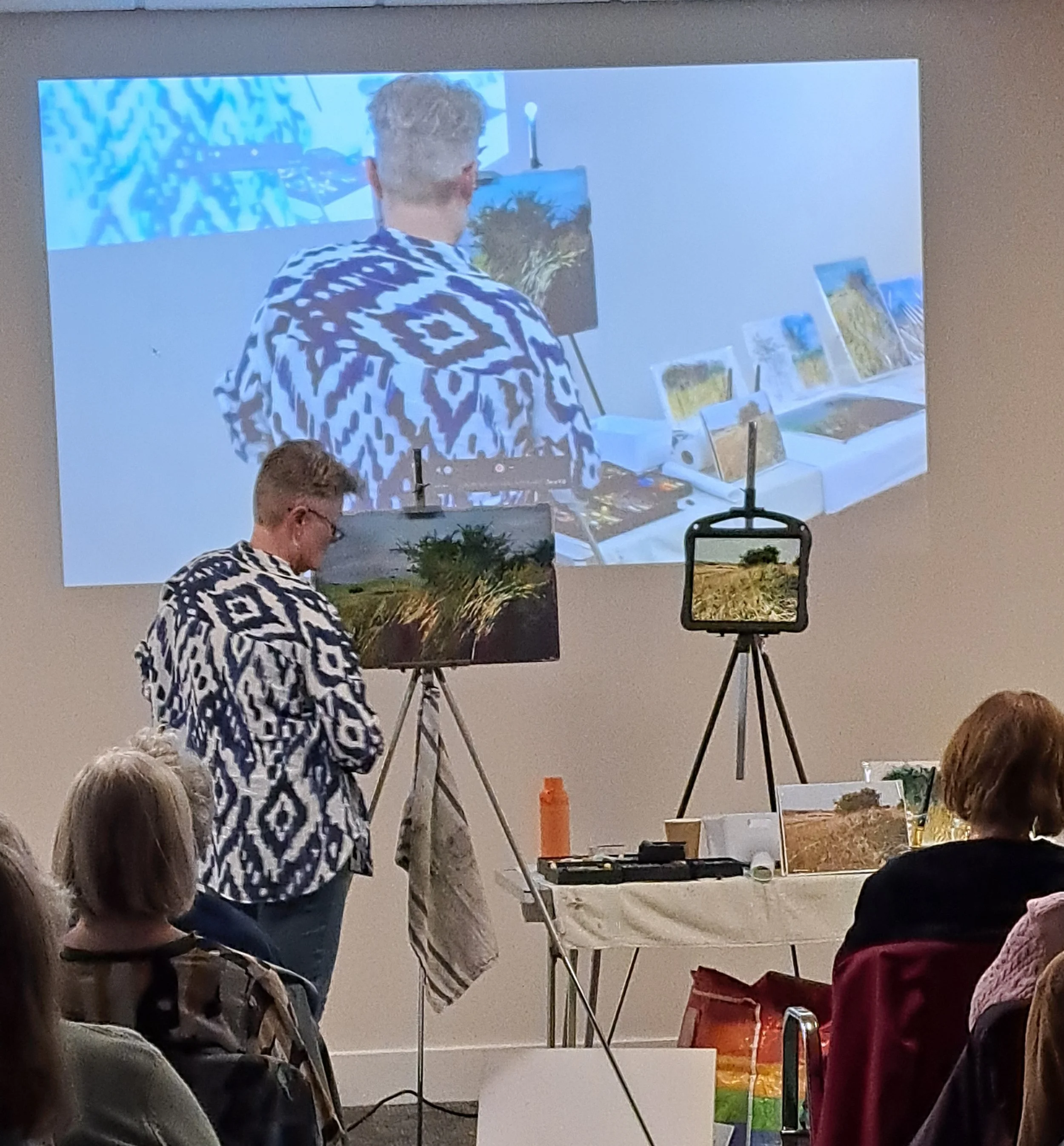 An artist in a patterned black and white shirt standing in front of easels displaying landscape paintings, with a large projection of the artist working on a painting on the wall behind.