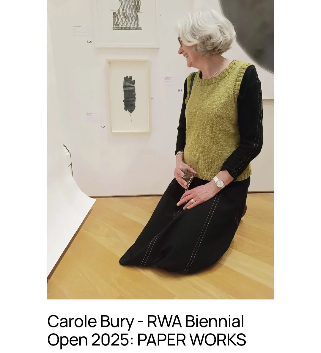 A woman with gray hair kneels on a wooden floor in an art gallery, holding a drink, looking at artwork on the wall.