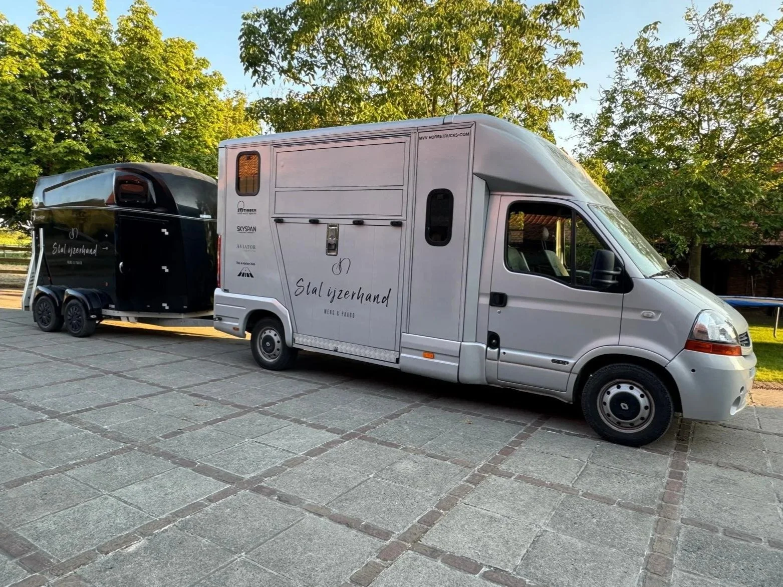 Een wit computervrachtwagen met een gesloten trailer die een zwarte caravan trekt, geparkeerd op een stenen plein met bomen in de achtergrond.