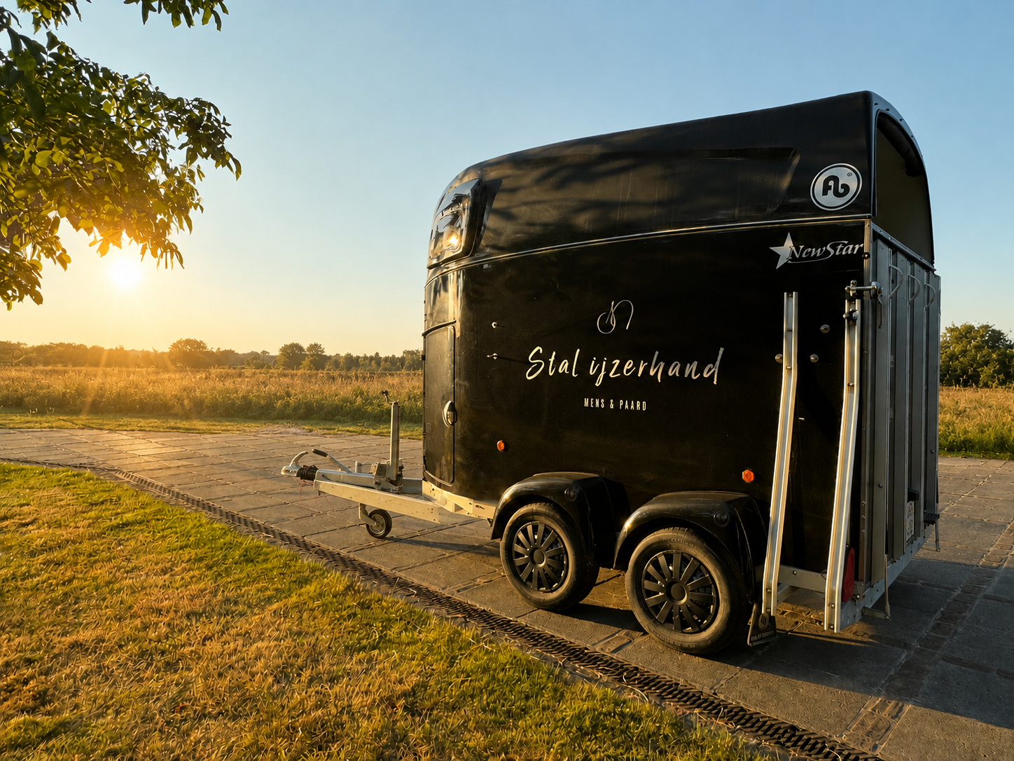 Een zwarte paardentrailer geparkeerd op een oprit bij zonsondergang, met de naam 'Stal ijzerhand' en de tekst 'MENS & PAARD' erop.