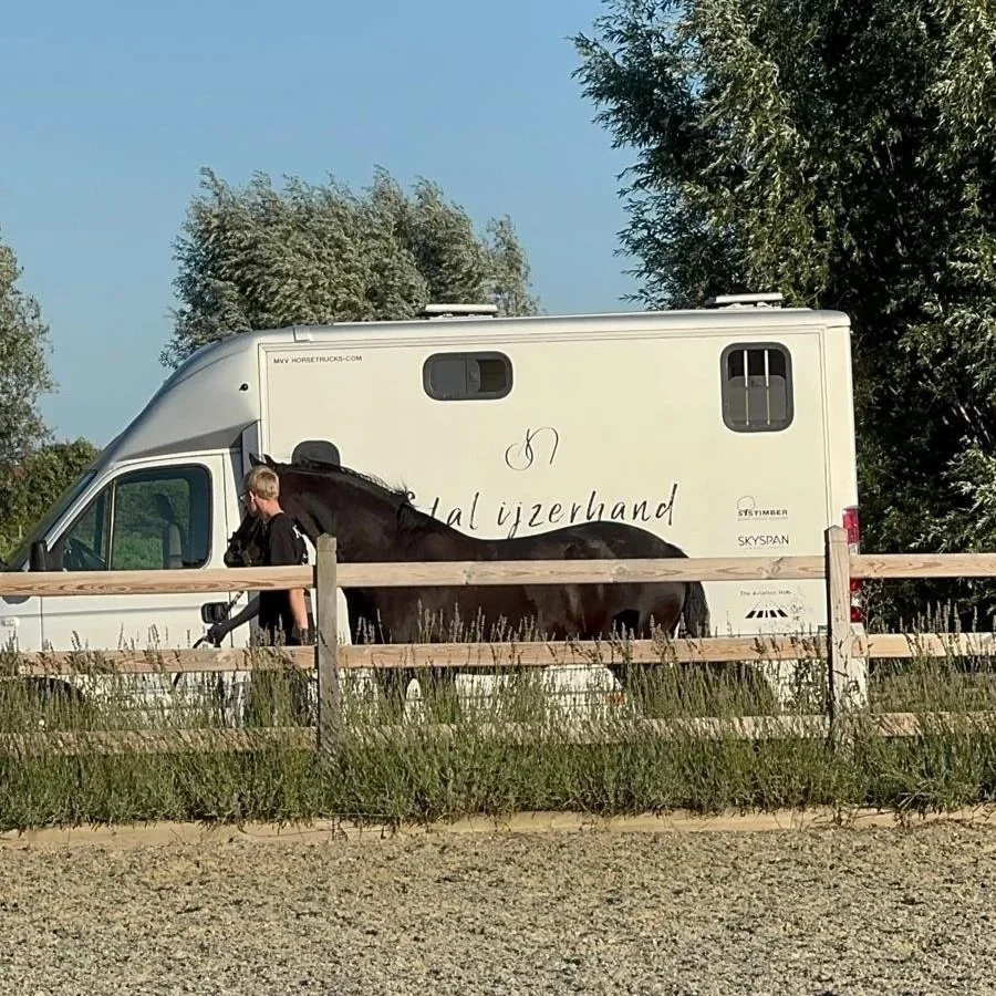 Een man met een zwart paard staat voor een witte camper op een omheinde weg met houten hek en groene bomen op de achtergrond.