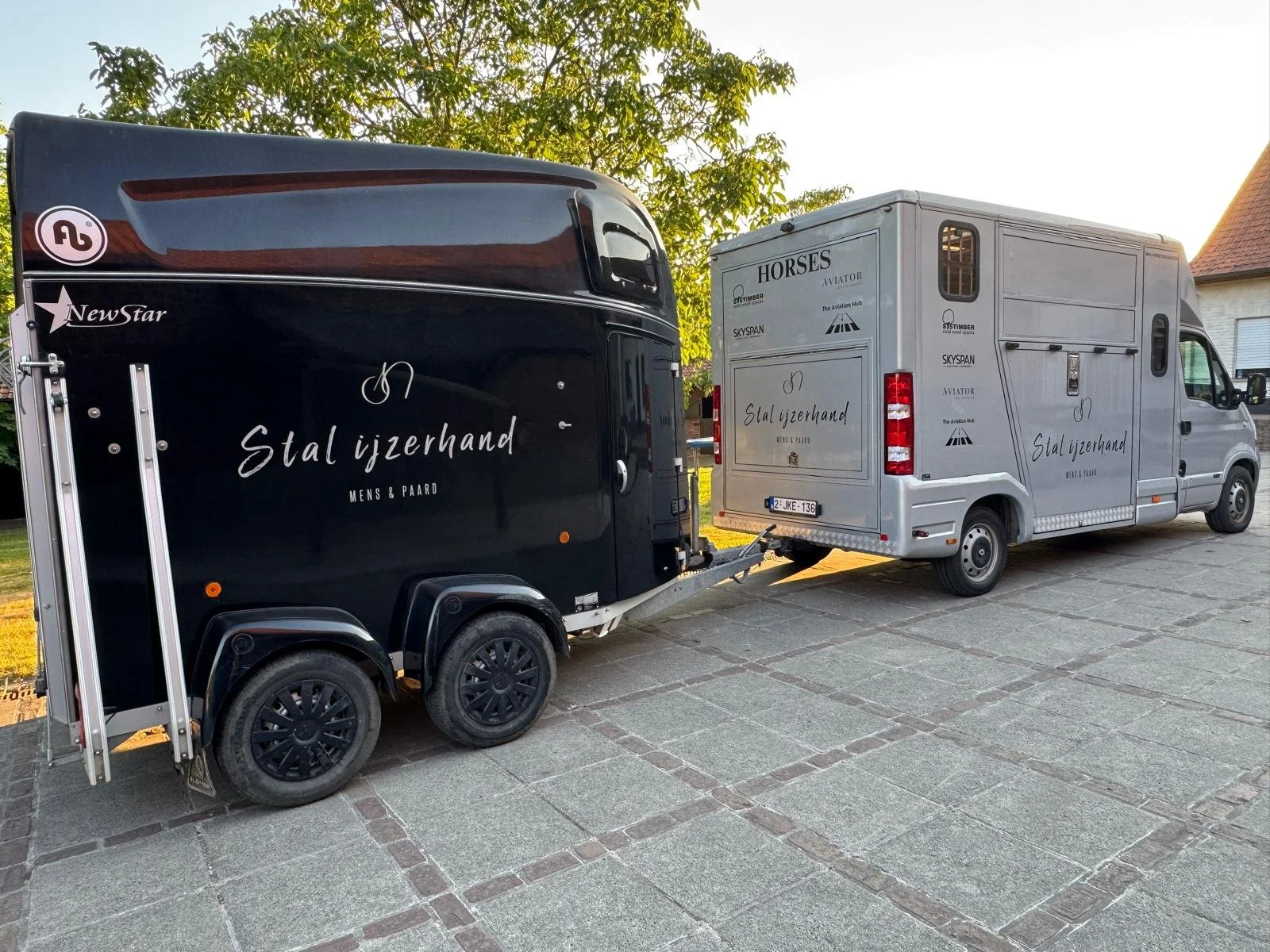 Een paardenwagen en een dubbeldeks bestelwagen geparkeerd op een stoep naast een huis, beide bedrukt met bedrijfsnaam 'Stal ijzerhand Mens & Paard'.