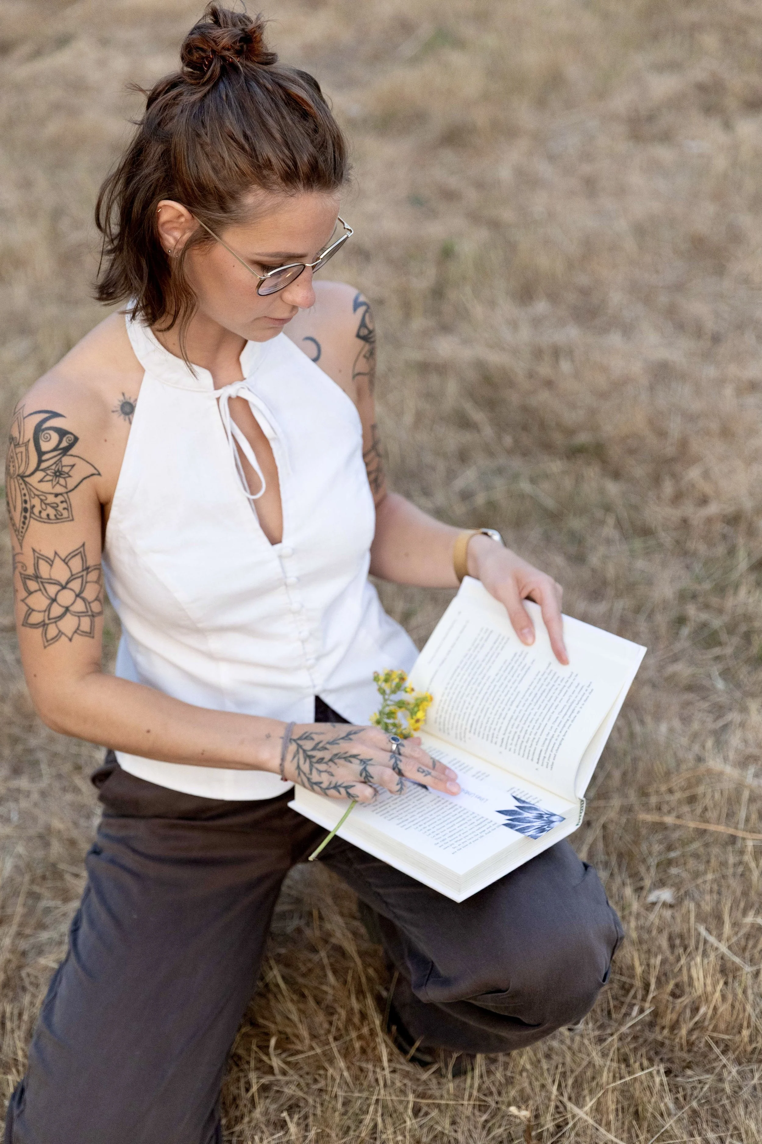 Een vrouw met tattoo's op haar armen die op de grond zit en een boek leest, met een veld op de achtergrond.