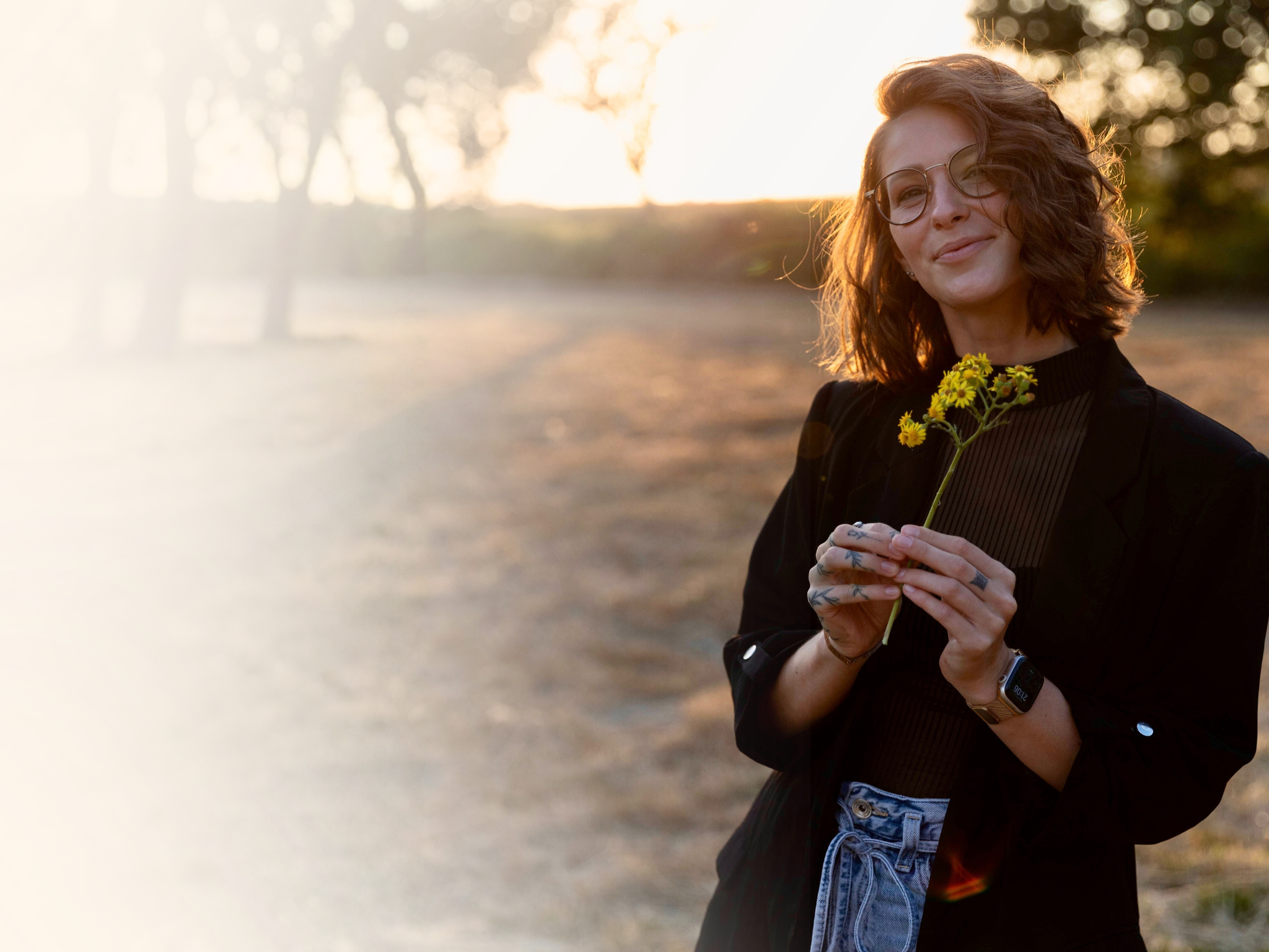 Vrouw met donkere krullen, bril en tattoos, die een gele bloemen op een bosje vasthoudt, tijdens zonsondergang buiten.