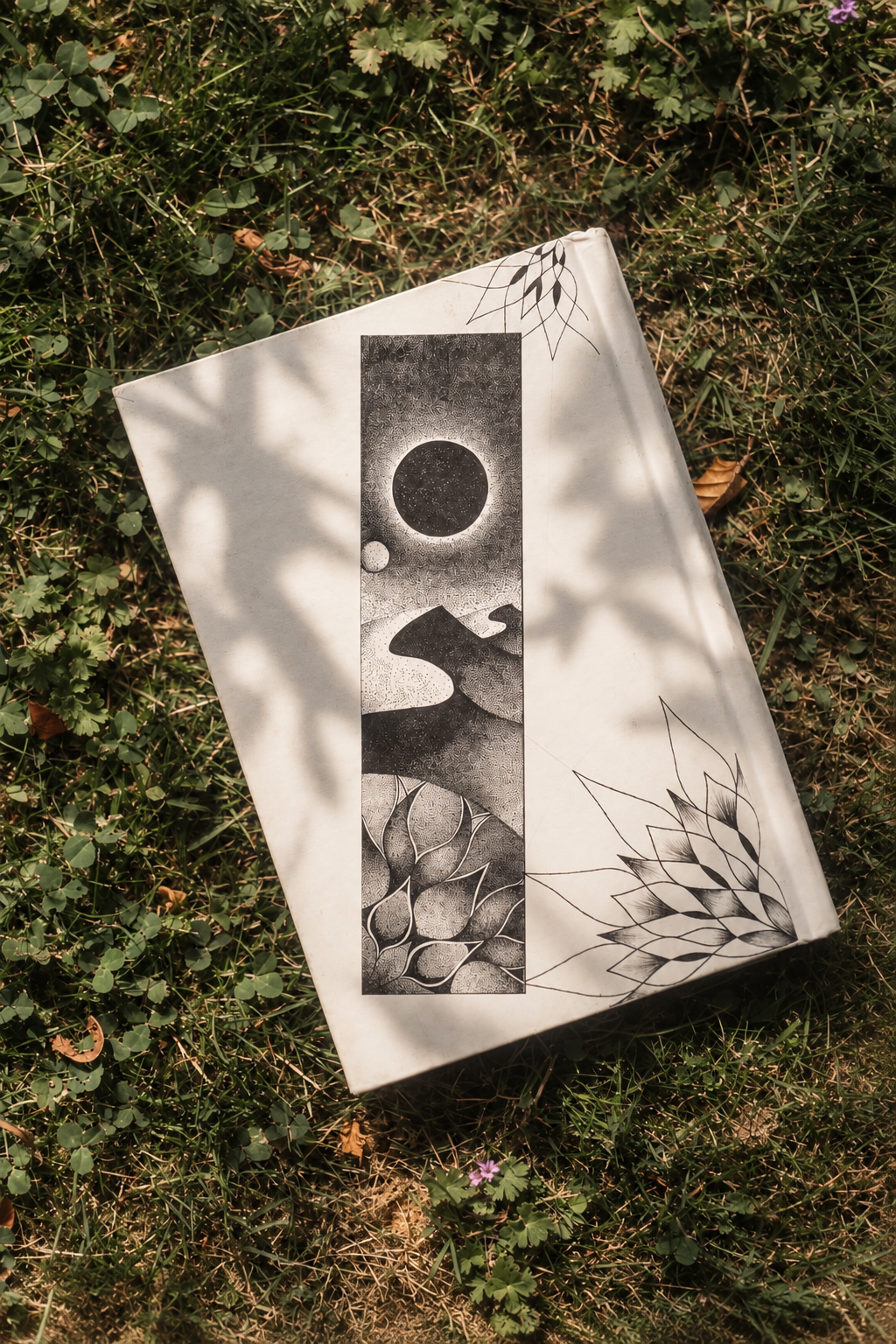 Een boek met een zwart-wit illustratie van een scène met een grote donkere cirkel en een kleinere licht cirkel, een berglandschap, en bloemachtige patronen, liggend op gras met kleine planten en een paar bladeren.