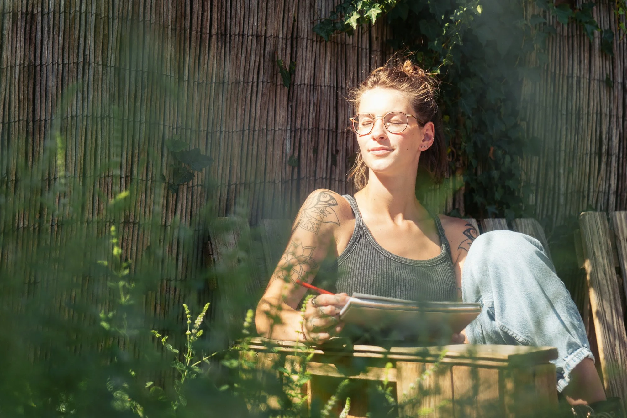 Een vrouw met korte bruin haar, oorbellen en een tatoeage op haar arm, zit buiten met haar ogen dicht in de zon, achter een houten schutting met groene klimplant.