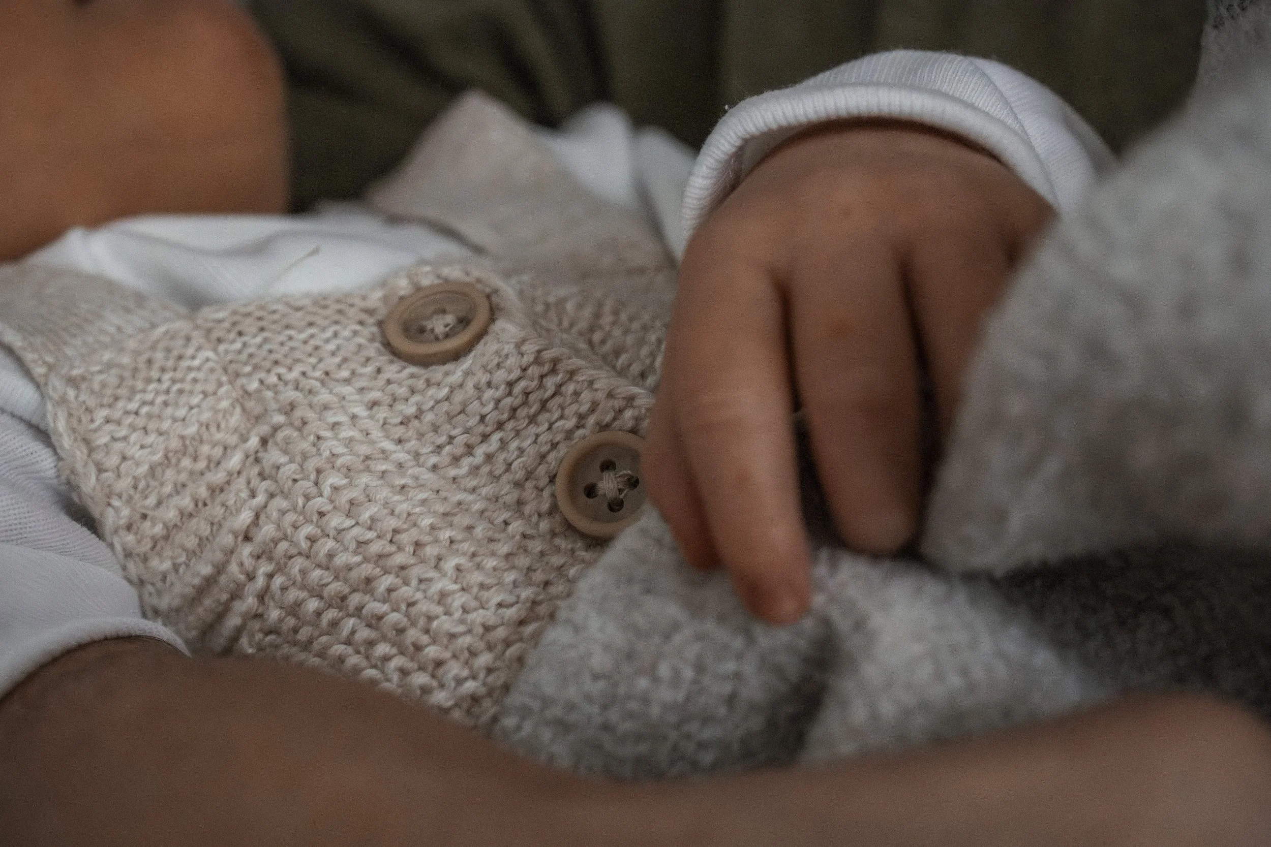 Nahaufnahme einer Babyhand, die eine graue Decke hält, getragen von einem Kind in grauer Strickjacke mit Knöpfen.