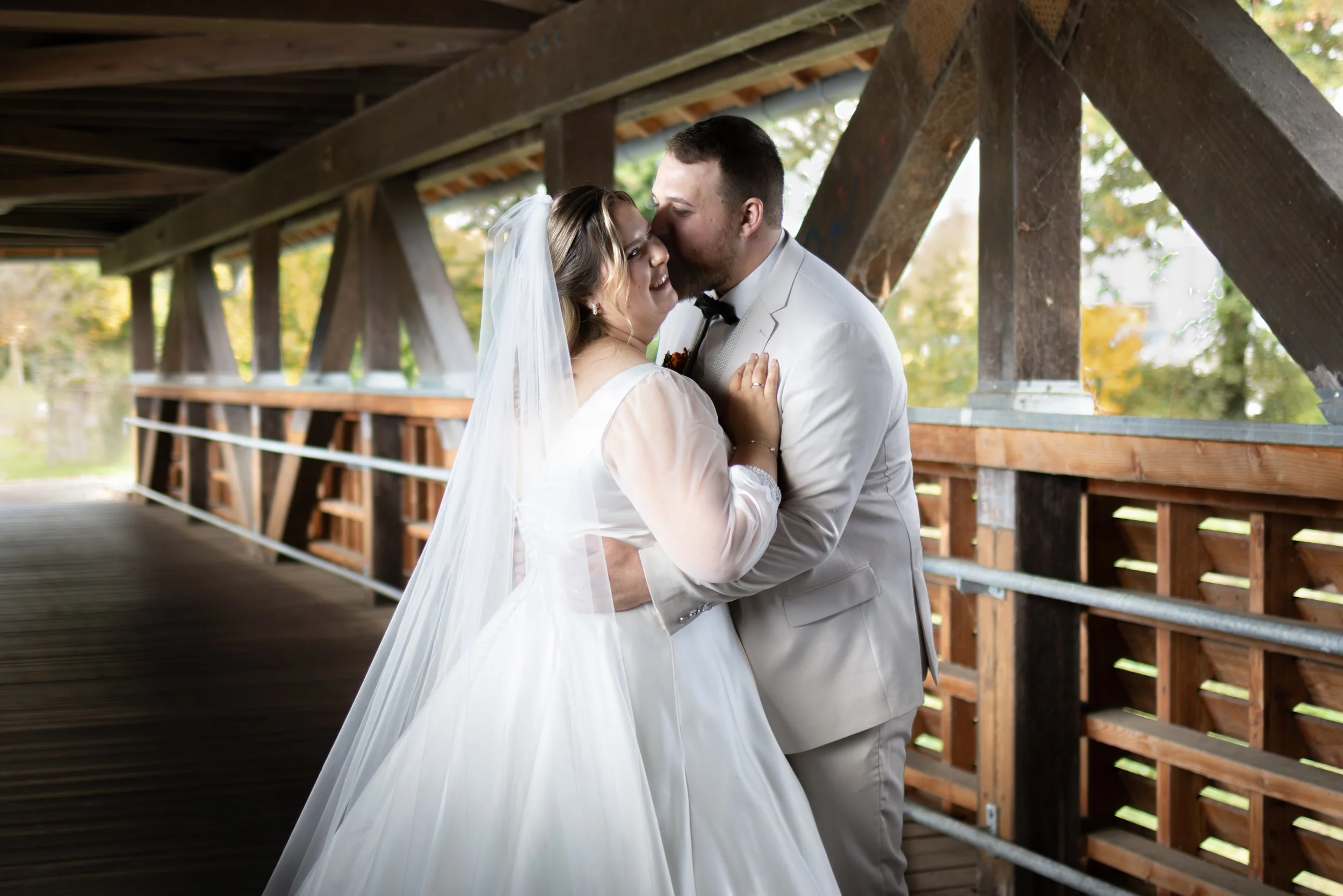 Ein Brautpaar in Hochzeitskleidung, das sich an einem Holzbrücke umarmt und küsst, im Freien mit herbstlichen Bäumen im Hintergrund.