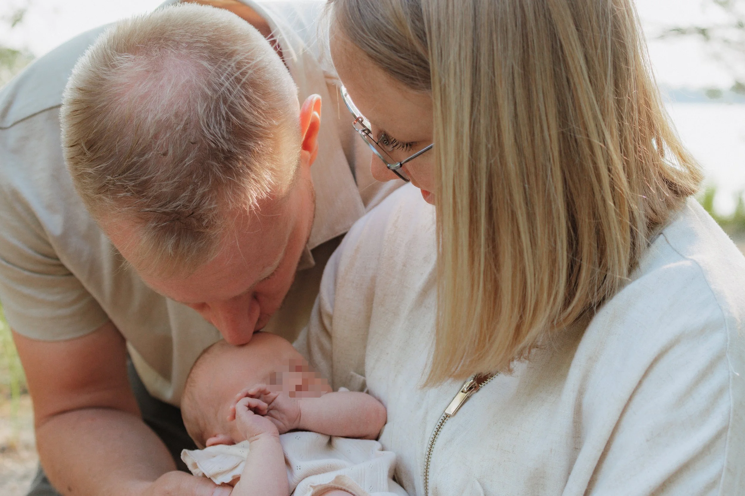 Ein Baby wird von einem Mann und einer Frau draußen gehalten. Der Mann taucht das Gesicht des Babys, die Frau hält das Baby, beide schauen liebevoll auf das Baby.