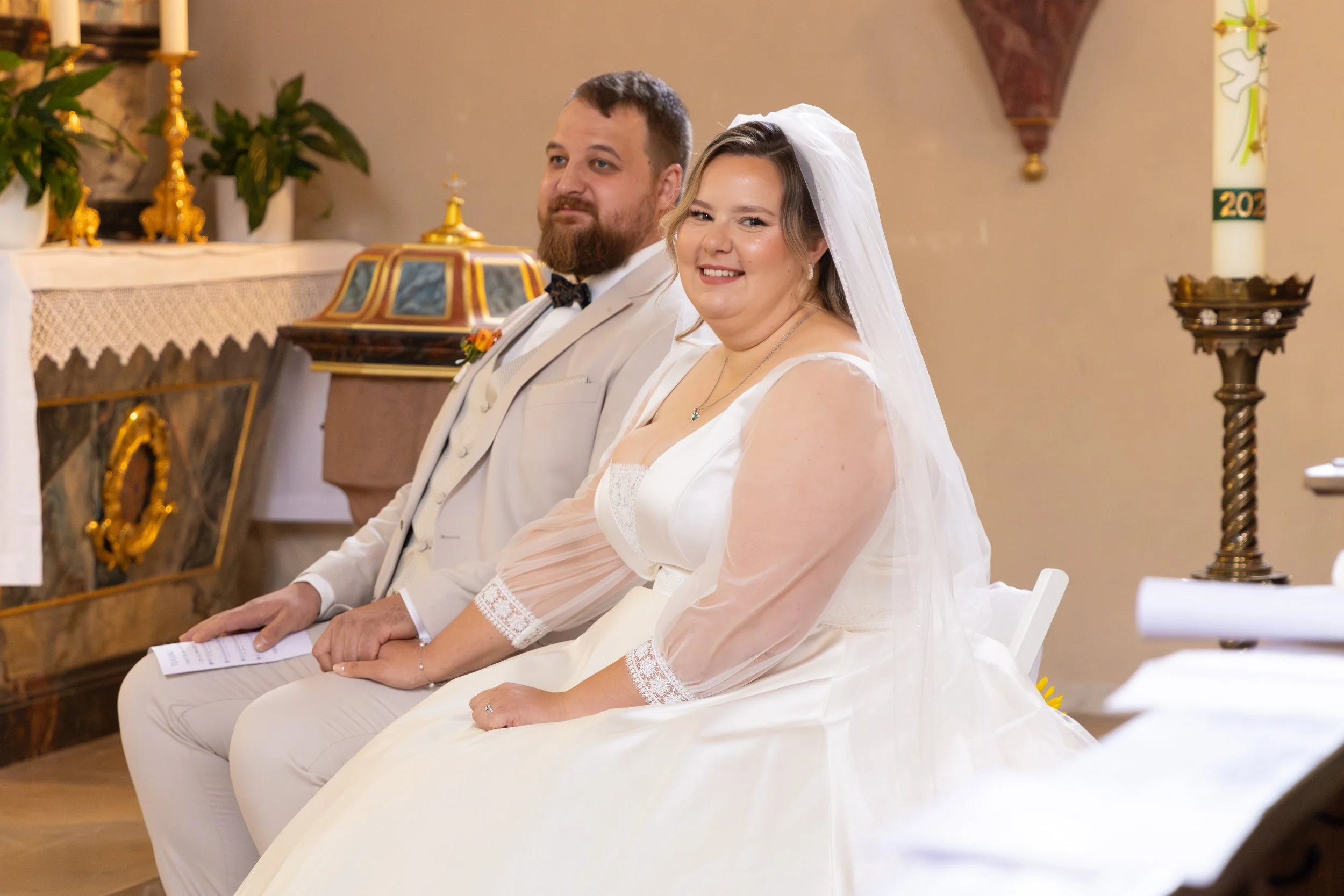 Ein verheiratetes Paar während einer Trauzeremonie in einer Kirche, beide sitzen nebeneinander, die Frau trägt ein weißes Hochzeitskleid mit Schleier, lächelt in die Kamera, der Mann trägt einen weißen Anzug mit Fliege.