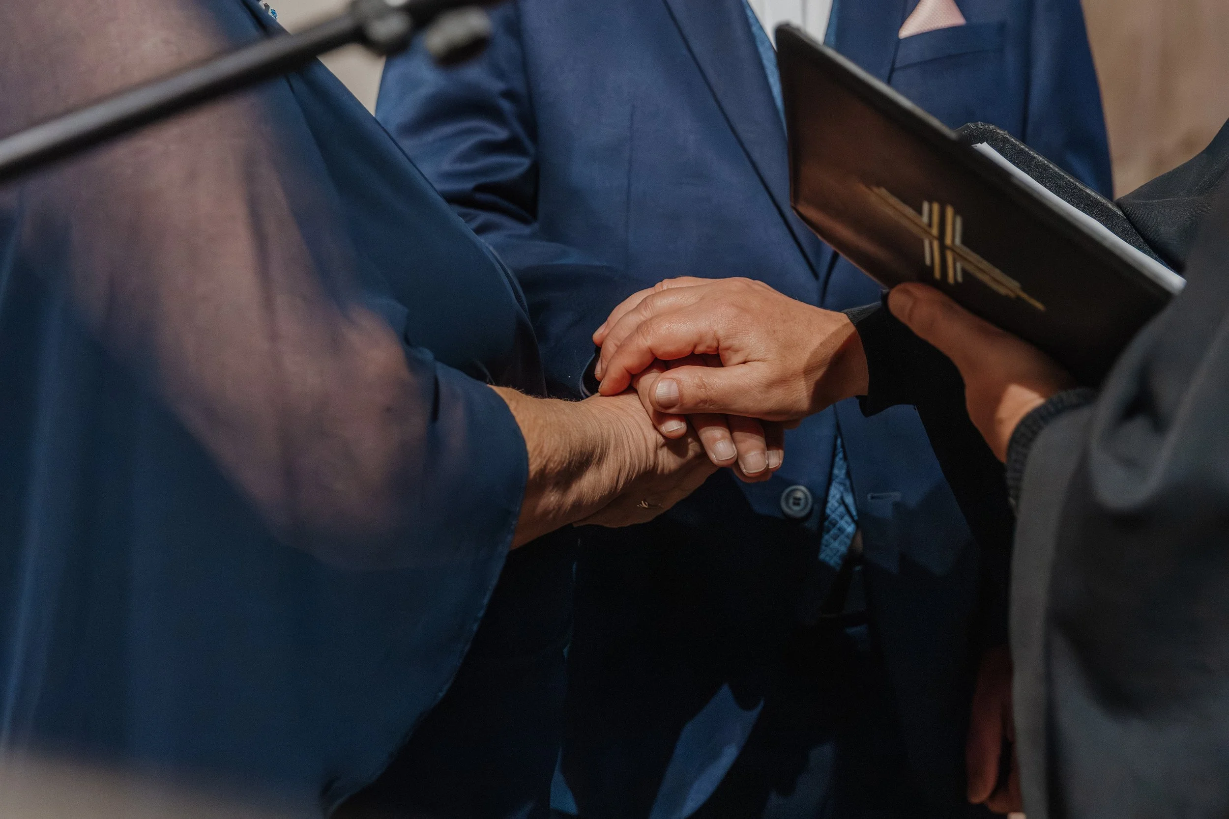 Eine Person hält die Hand einer anderen Person in einer religiösen Zeremonie, vermutlich bei einer Heirat oder Taufe, während eine dritte Person ein Buch hält.